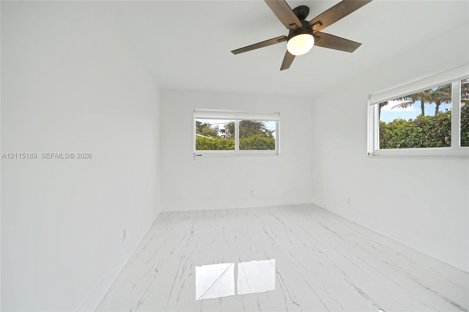 Light-filled interior featuring stone-finish flooring with veining, dual windows, and a dark-finish ceiling fan with integrated lighting