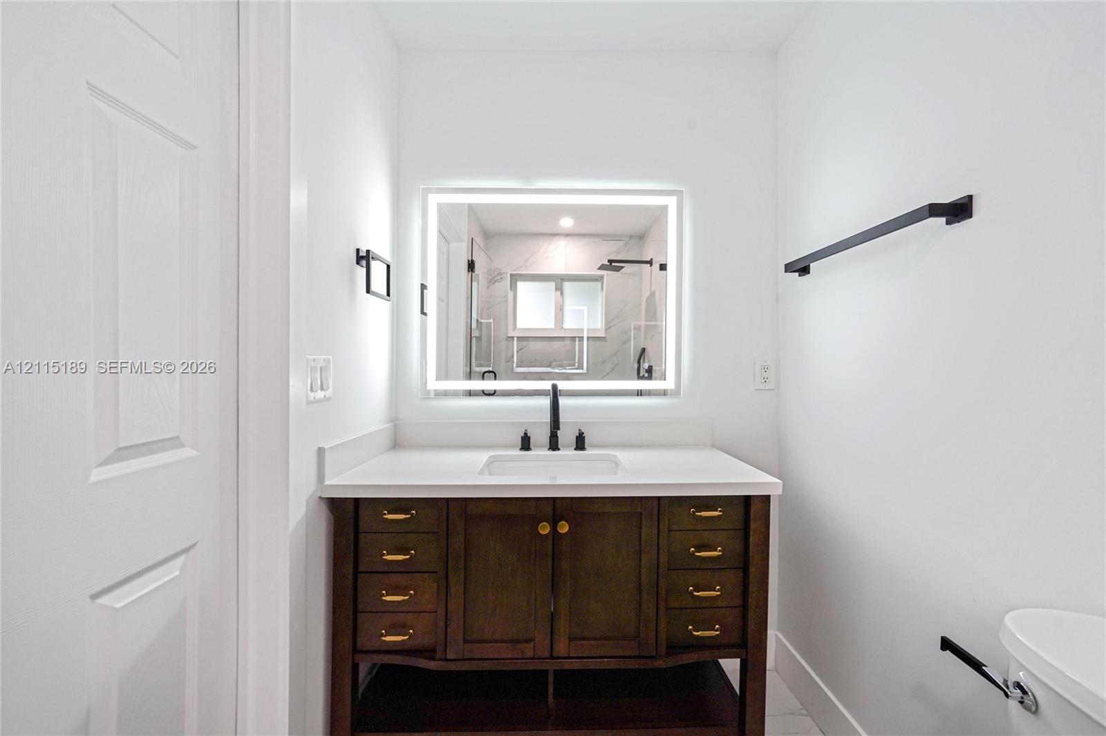 Bathroom vanity with a dark wood finish, white countertop with an integrated rectangular sink, matte black faucet, and backlit wall-mounted mirror, accented by matte black towel bars