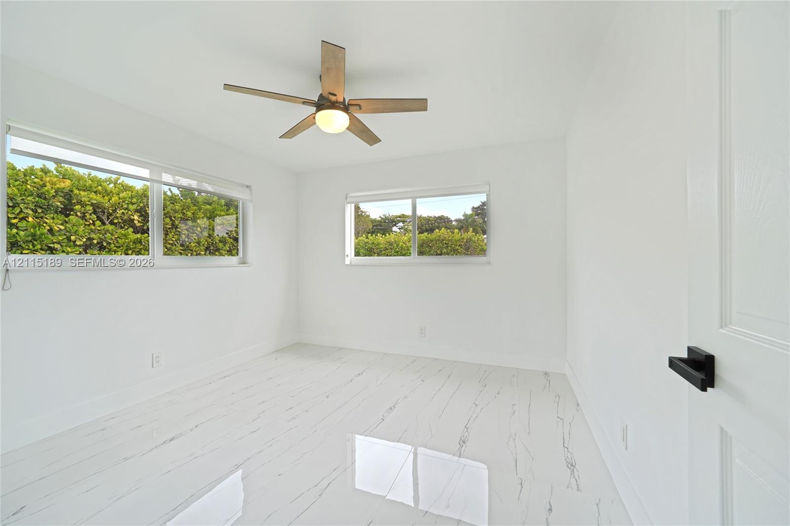 Room featuring polished white tile flooring with veining, large horizontal windows, a wooden-finish ceiling fan with integrated lighting, and a door with matte black hardware