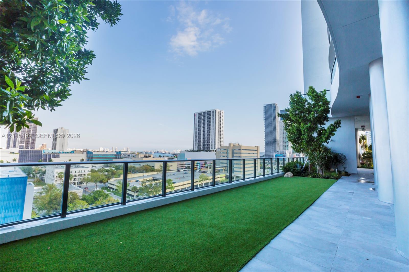 Covered terrace featuring large-format tile flooring, artificial turf, structural columns, and a glass-paneled railing