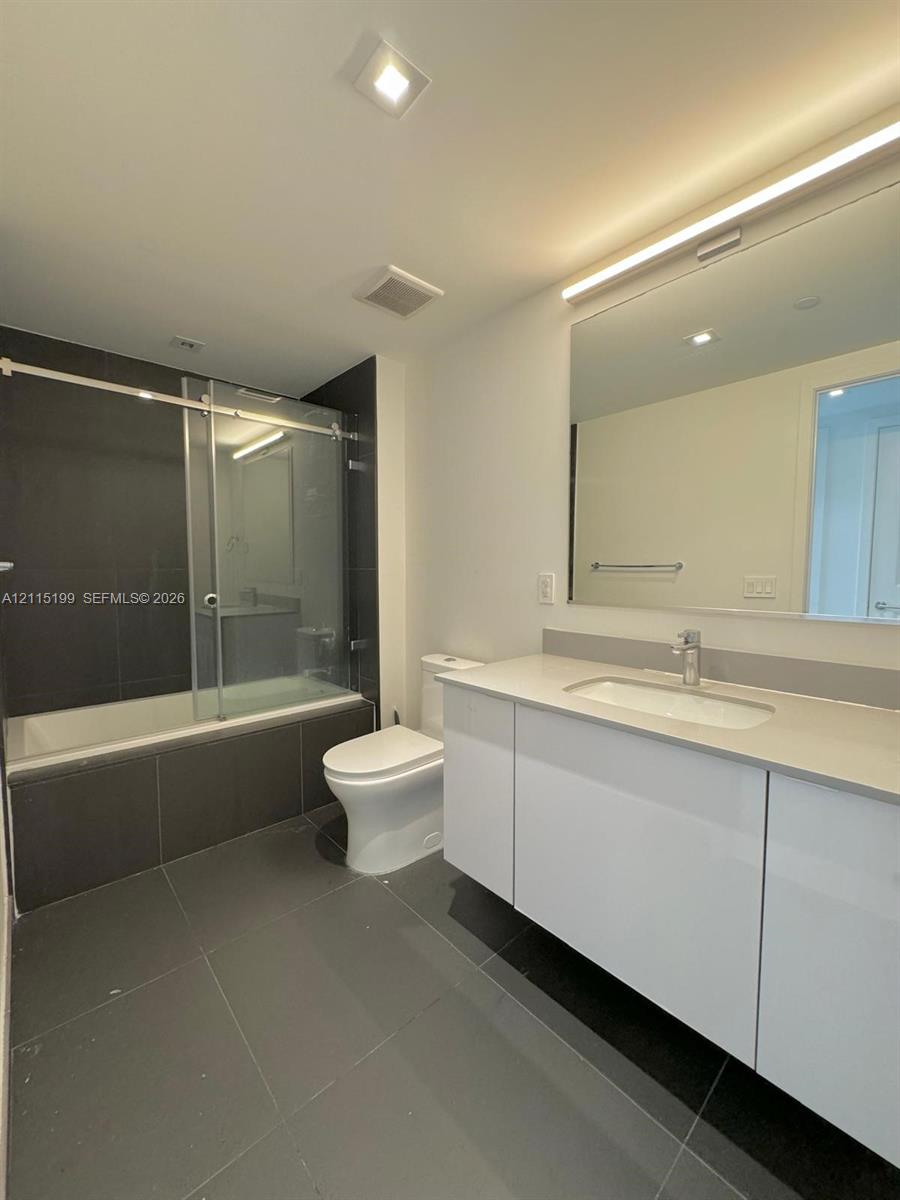 Bathroom featuring a white vanity with a neutral solid-surface countertop, an integrated undermount sink, a large wall-mounted mirror with integrated lighting, a soaking tub with a glass sliding enclosure, and large-format gray tile flooring