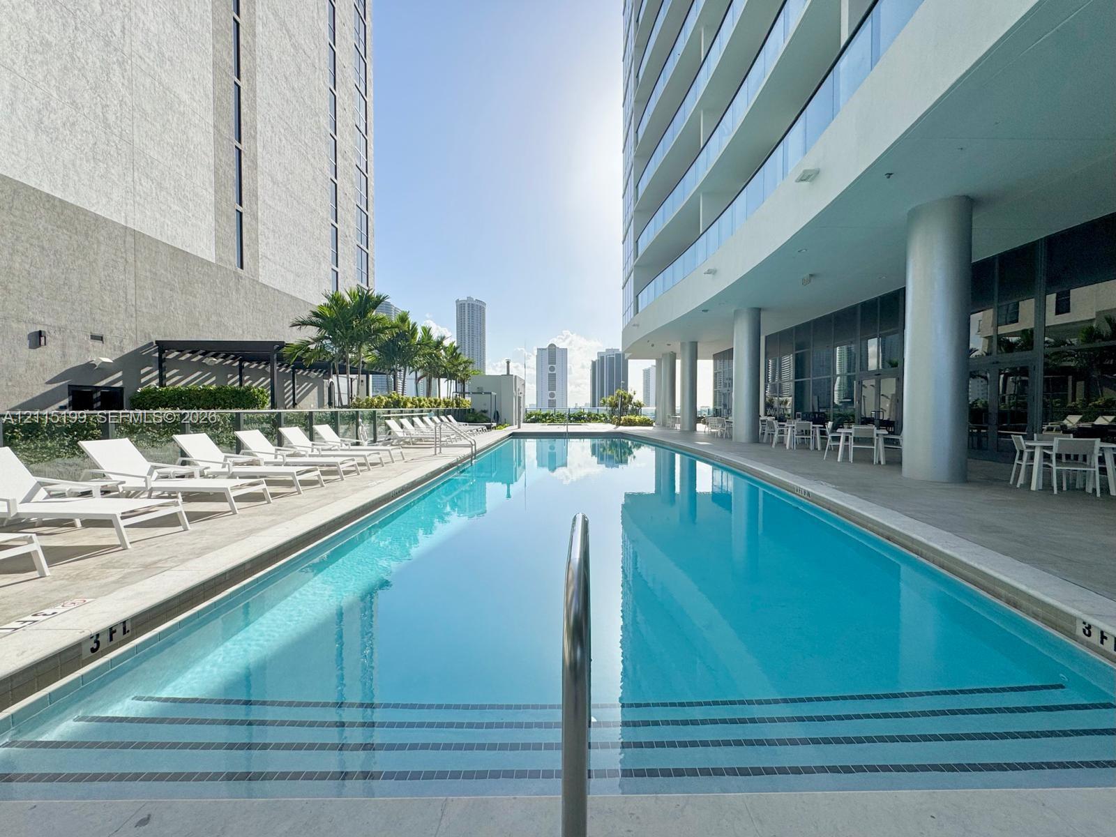 Rectangular swimming pool with tiled entry steps, expansive stone-paved pool deck, glass balcony railings, and structural columns