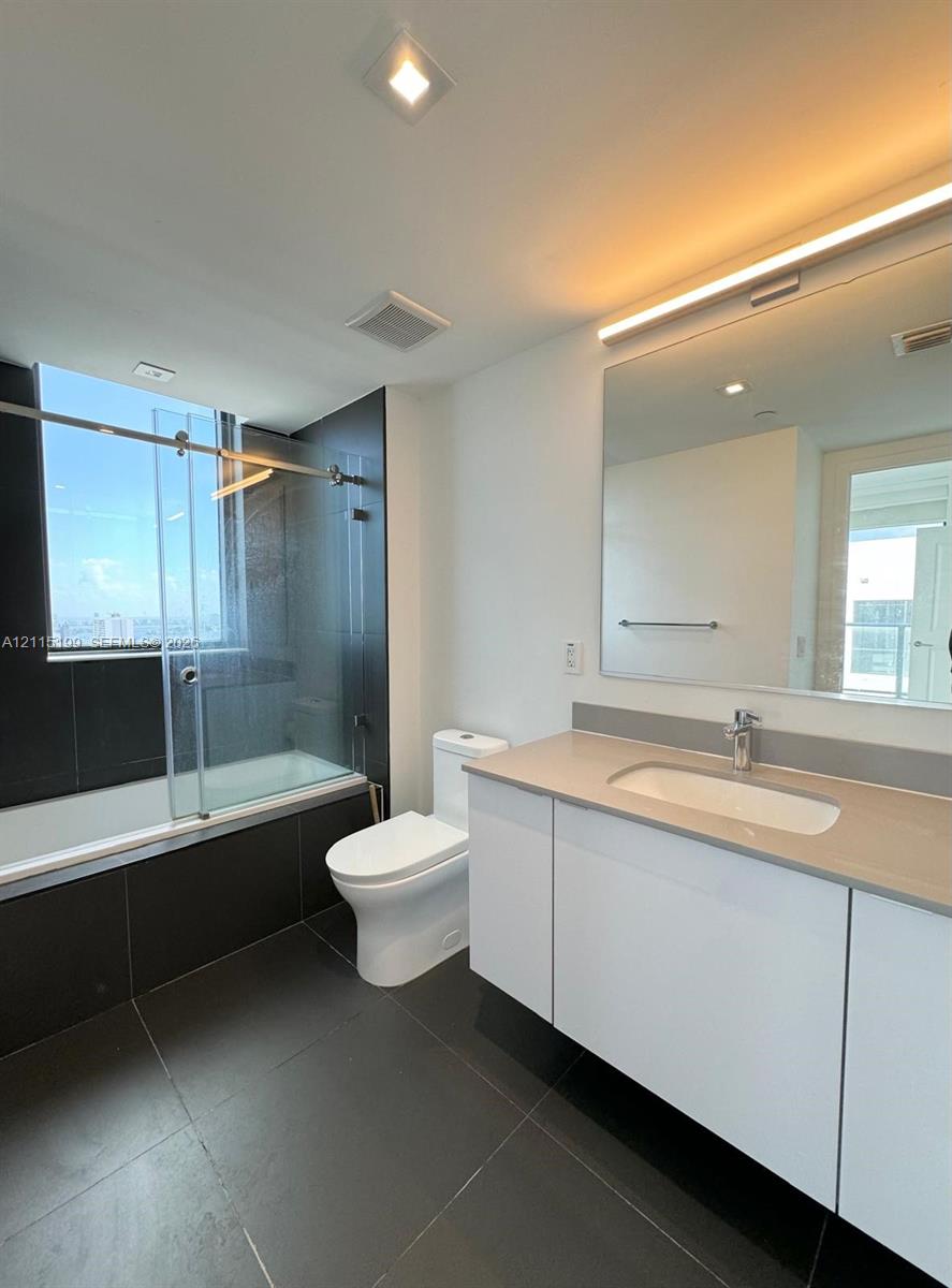 Bathroom featuring a white flat-panel vanity with an integrated countertop, large wall-mounted mirror with integrated lighting, oversized charcoal floor tile, and a soaking tub with a sliding glass enclosure and charcoal tile surround