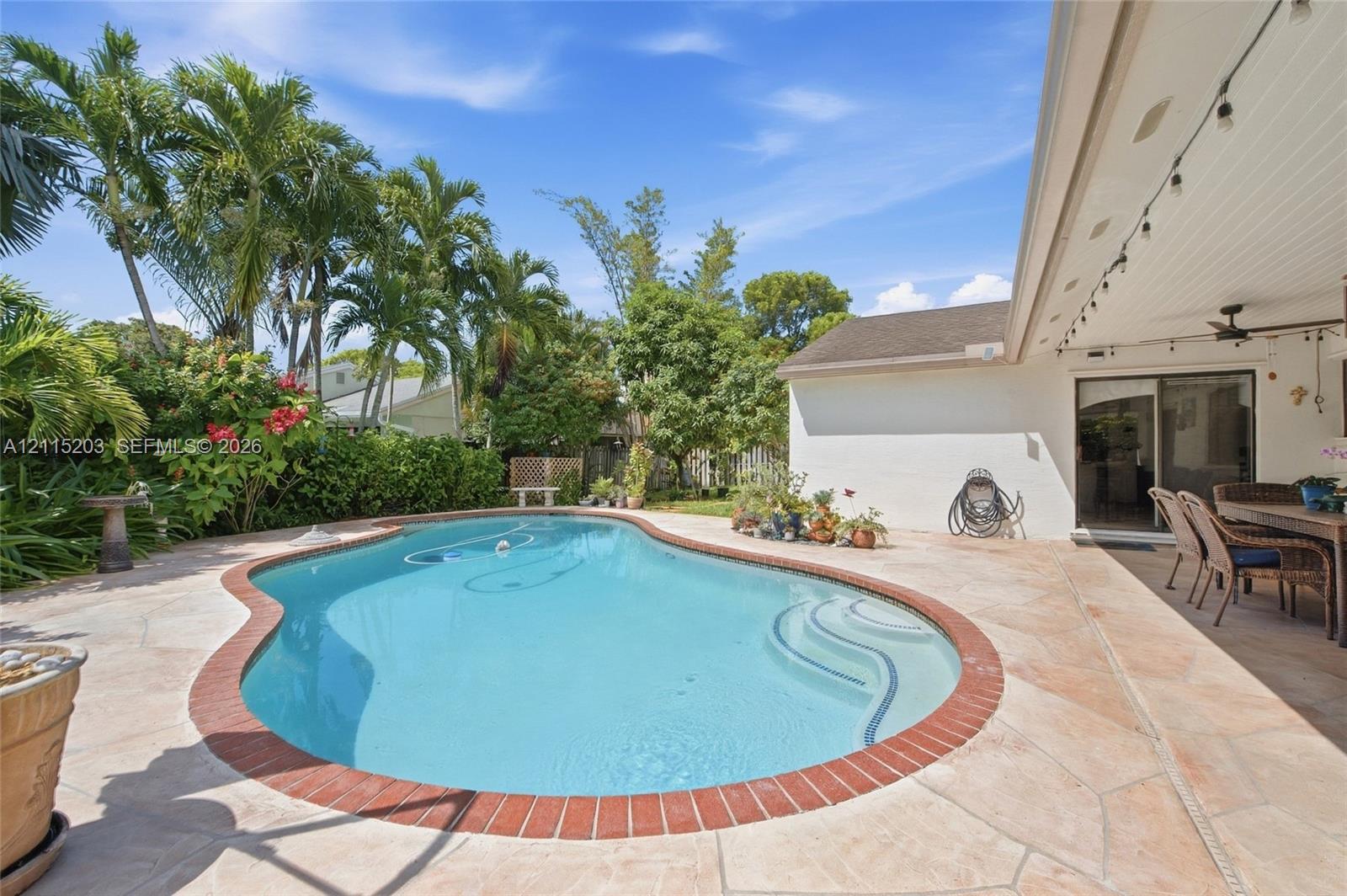 Free-form swimming pool with integrated steps, surrounded by natural stone pavers and a brick coping border, featuring a covered patio and tropical landscaping Free-form swimming pool with integrated steps, surrounded by natural stone pavers and a brick coping border, featuring a covered patio and tropical landscaping