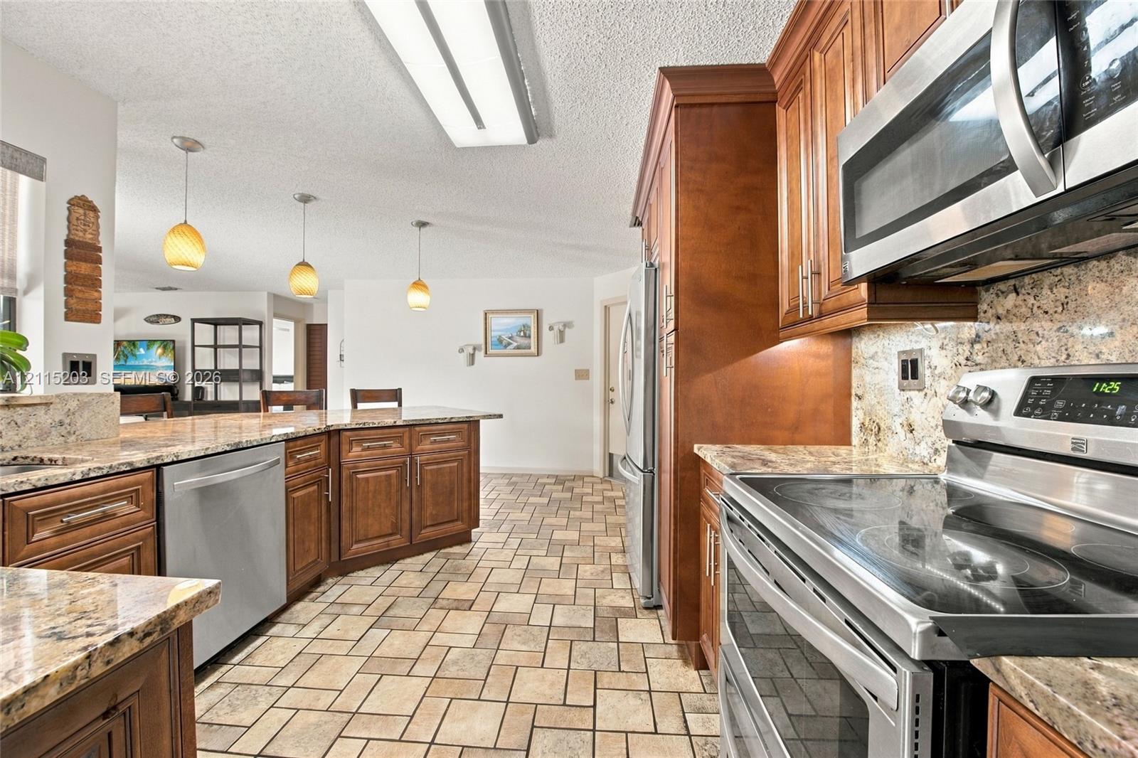 Kitchen featuring stained wood cabinetry, stone-finish countertops, stainless steel appliances, ceramic tile flooring, and three pendant light fixtures