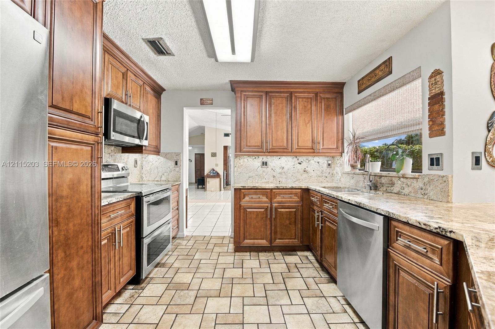 Kitchen featuring warm-tone wood cabinetry, natural stone countertops, stainless steel appliances, and tiled flooring