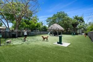 Fenced outdoor recreation area featuring synthetic turf, a thatched-roof pavilion, mature trees, and a water station