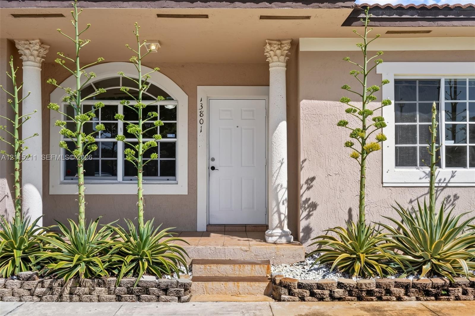 Stucco exterior featuring dual decorative columns with ornate capitals, an arched window, and a stone-trimmed garden bed