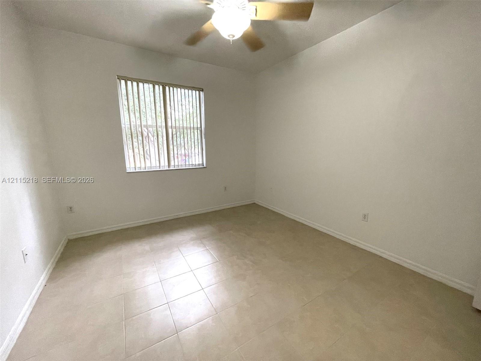 Neutral-toned tile flooring, light-colored wall finish, ceiling-mounted fan with integrated lighting, and a single window featuring vertical blinds