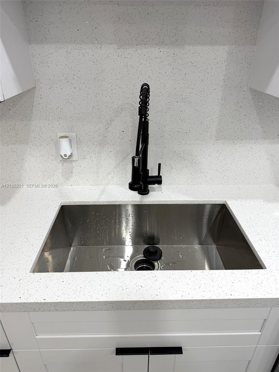 Undermount stainless steel sink featuring a high-arc matte black pull-down faucet, white flecked stone-composite countertop, matching full-height backsplash, and shaker-style cabinetry with matte black hardware