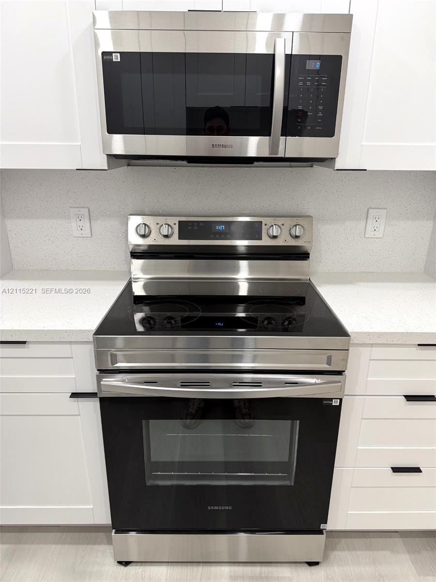Stainless steel electric range with smooth-top cooktop and matching over-the-range microwave, set between white shaker-style cabinetry, complemented by solid-surface countertops