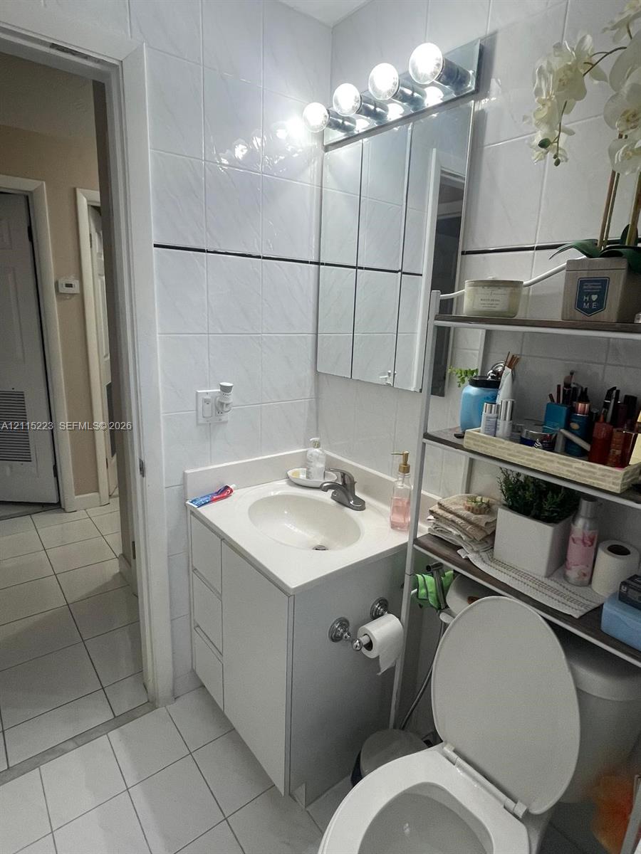 Bathroom featuring white tiled walls with a horizontal decorative accent, a vanity with a white countertop and integrated sink, a multi-bulb vanity light fixture, and floor tiles