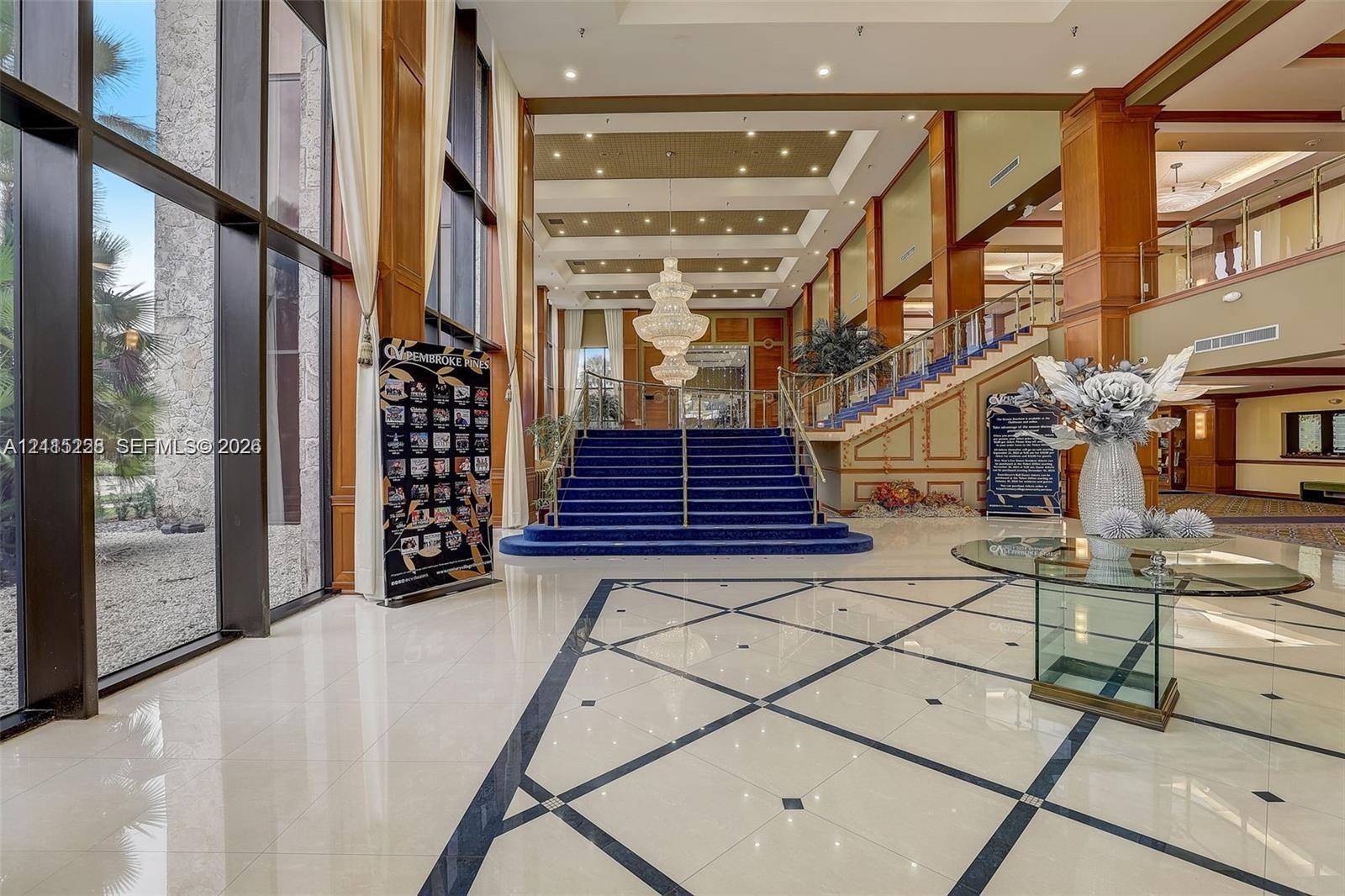 Grand lobby featuring floor-to-ceiling windows, polished tile flooring with inlaid geometric detailing, a tiered crystal chandelier, and a grand staircase with metal railings