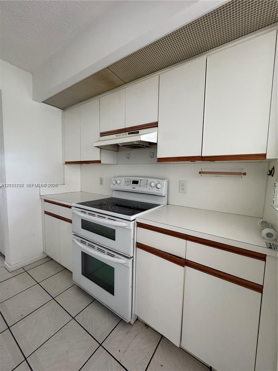 Kitchen featuring white cabinetry with wood trim, solid-surface countertops, a double-oven range with a glass cooktop, an integrated range hood, and tile flooring