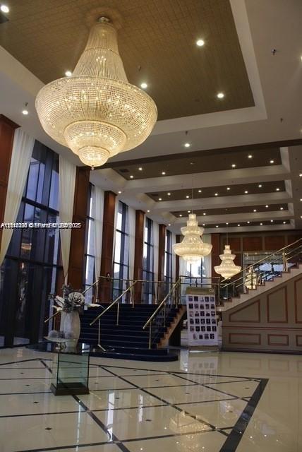 Grand lobby featuring multi-tiered crystal chandeliers, coffered ceilings with recessed lighting, floor-to-ceiling windows, polished stone flooring with decorative inlay, and an open staircase with metal railing