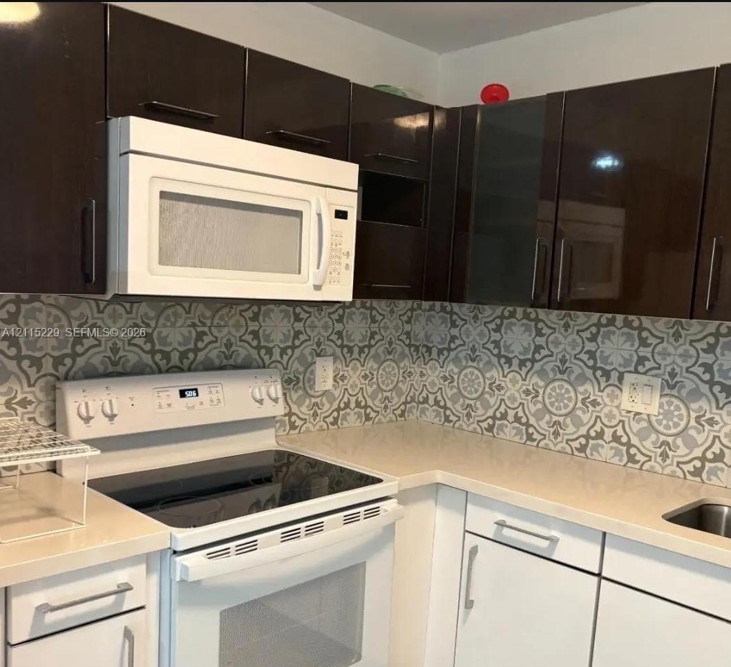 Kitchen featuring dark-toned upper cabinetry, white lower cabinetry with modern hardware, patterned tile backsplash, solid-surface countertops, and a white over-the-range microwave