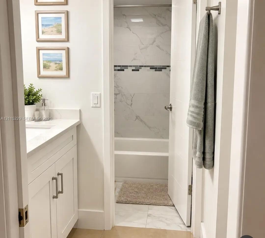White shaker-style vanity with a light-toned solid surface countertop and undermount sink, complemented by large-format light-toned tile flooring and a tub-shower enclosure featuring decorative mosaic tile accents