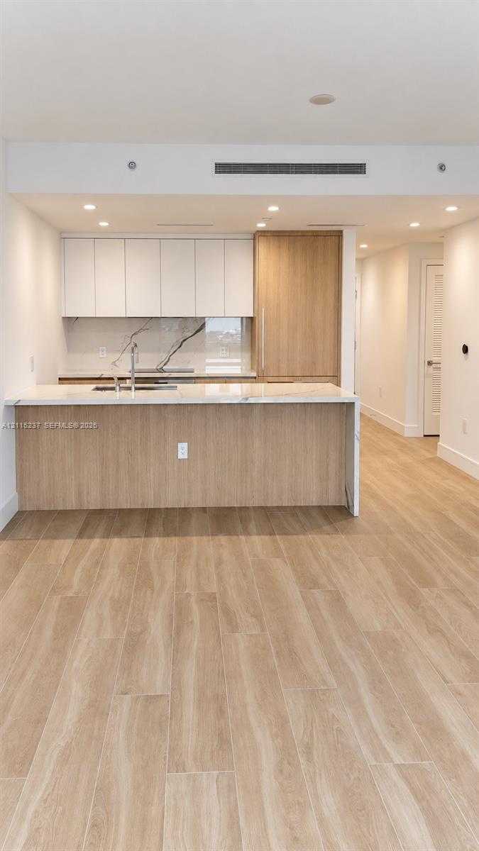 Kitchen featuring flat-panel cabinetry, light-tone wood-finish island, stone-surface countertops, stone-patterned backsplash, and wood-finish flooring