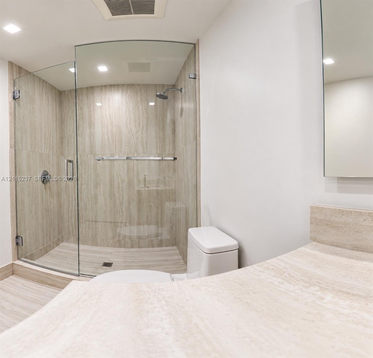 Walk-in shower with a curved glass enclosure and stone-finish wall tile, complemented by a stone-finish vanity countertop and a wall-mounted mirror