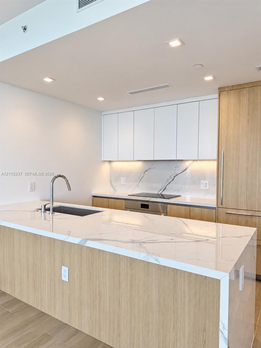 Kitchen featuring a waterfall-edge stone countertop, wood-finish cabinetry, integrated appliances, under-cabinet lighting, and a stone slab backsplash Kitchen featuring a waterfall-edge stone countertop, wood-finish cabinetry, integrated appliances, under-cabinet lighting, and a stone slab backsplash