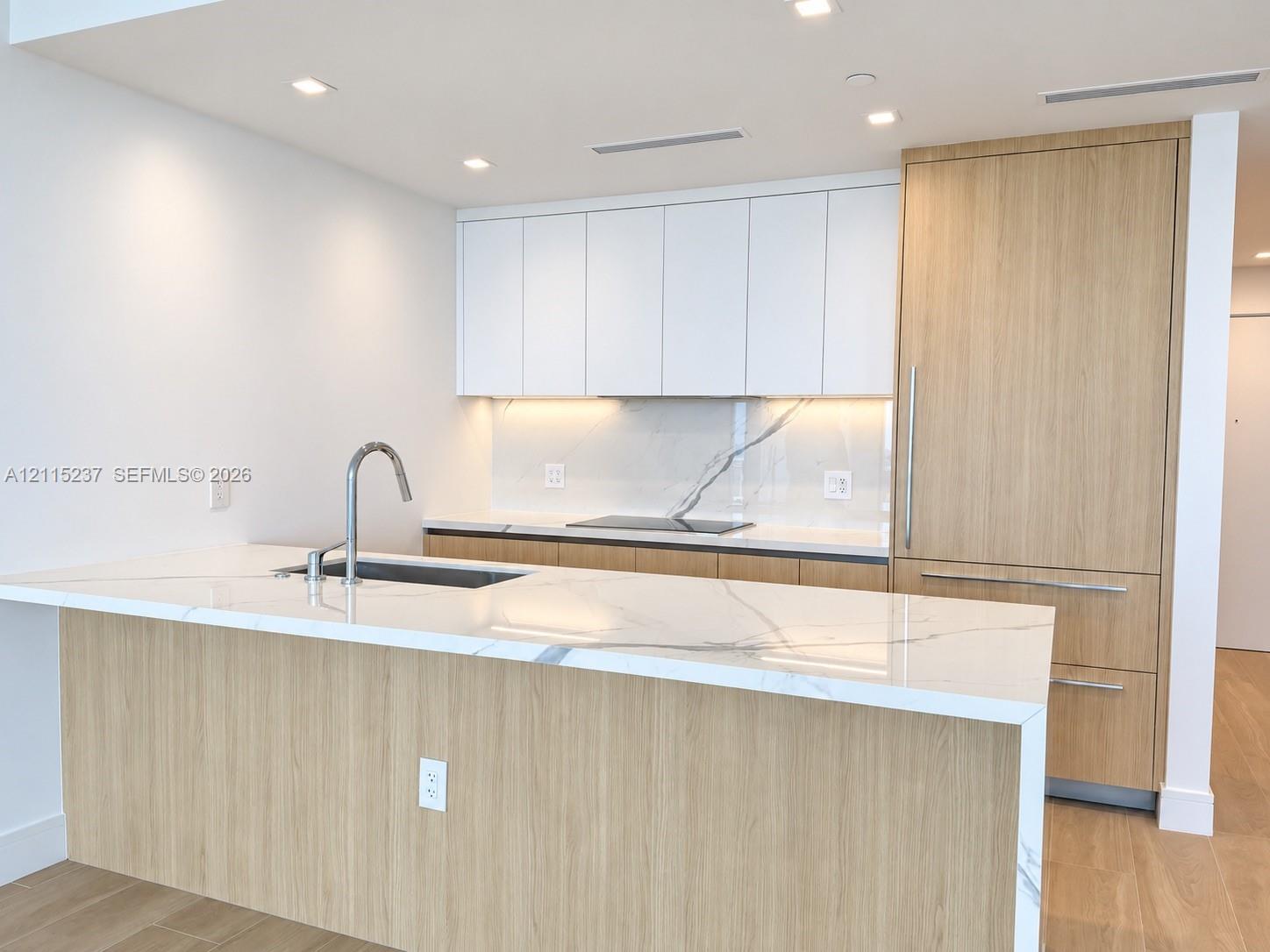 Kitchen featuring a waterfall-edge island with stone-finish countertop, flat-panel cabinetry with wood-finish and matte white cabinetry, integrated refrigerator, and stainless steel gooseneck faucet Kitchen featuring a waterfall-edge island with stone-finish countertop, flat-panel cabinetry with wood-finish and matte white cabinetry, integrated refrigerator, and stainless steel gooseneck faucet