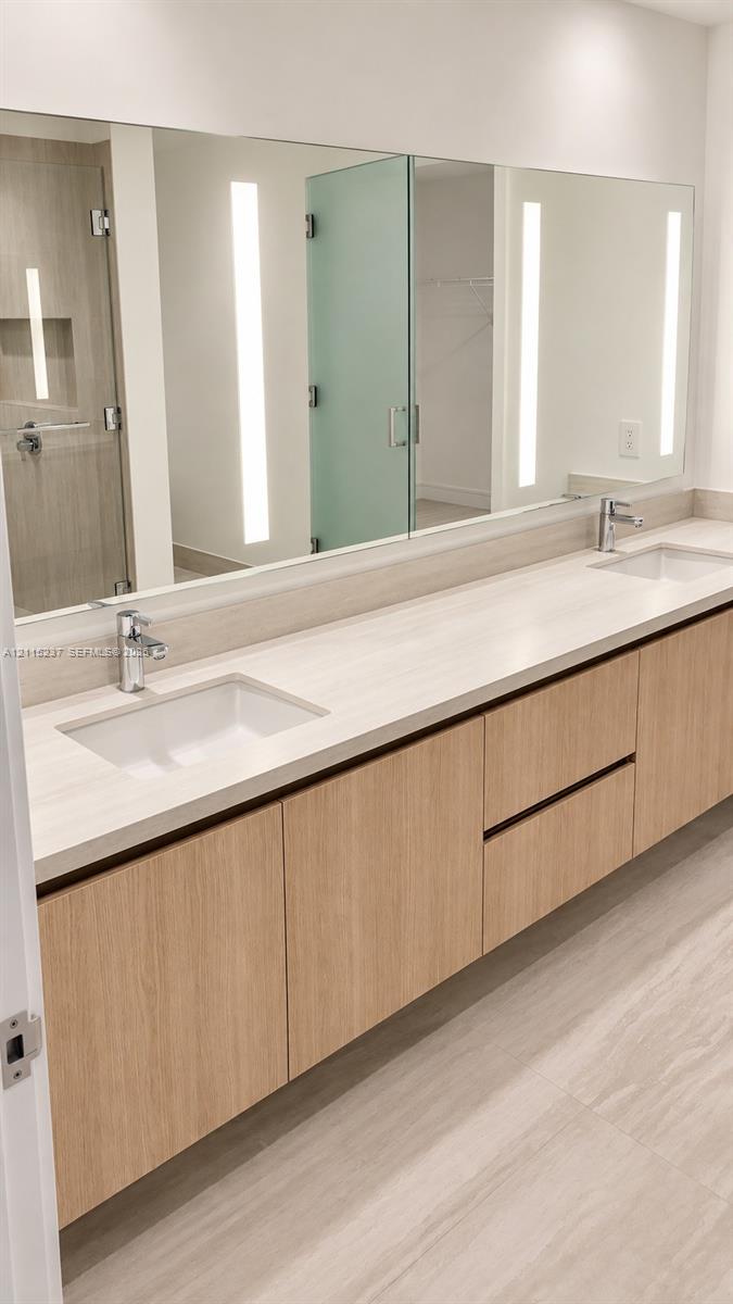 Floating wood-finish vanity with dual integrated rectangular sinks, light-toned stone-finish countertop, integrated linear vertical LED mirror lighting, and polished chrome fixtures