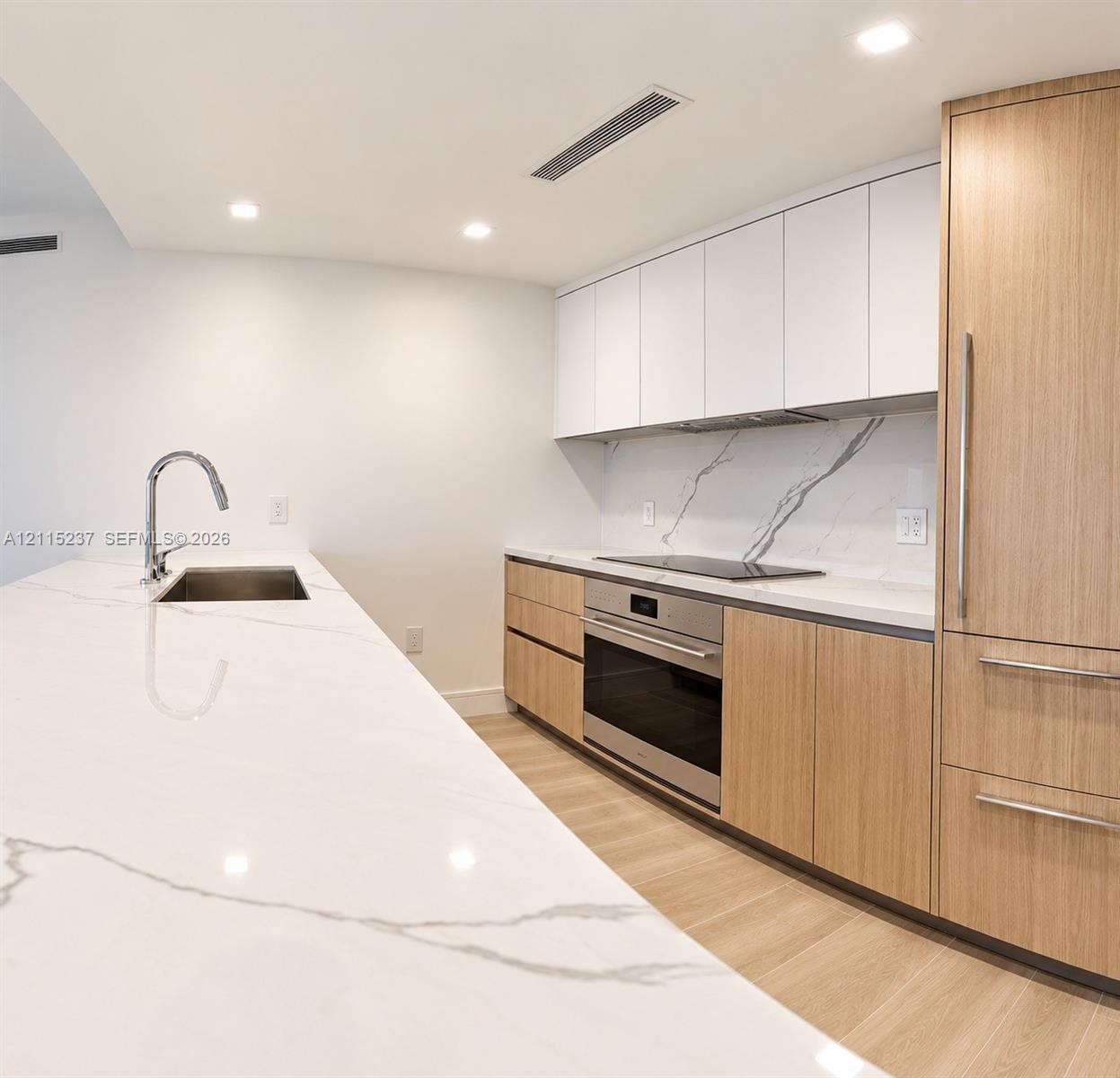 Kitchen featuring an expansive stone countertop with integrated sink, wood-finish cabinetry, white matte upper cabinets, stone backsplash with veining, and stainless steel built-in appliances