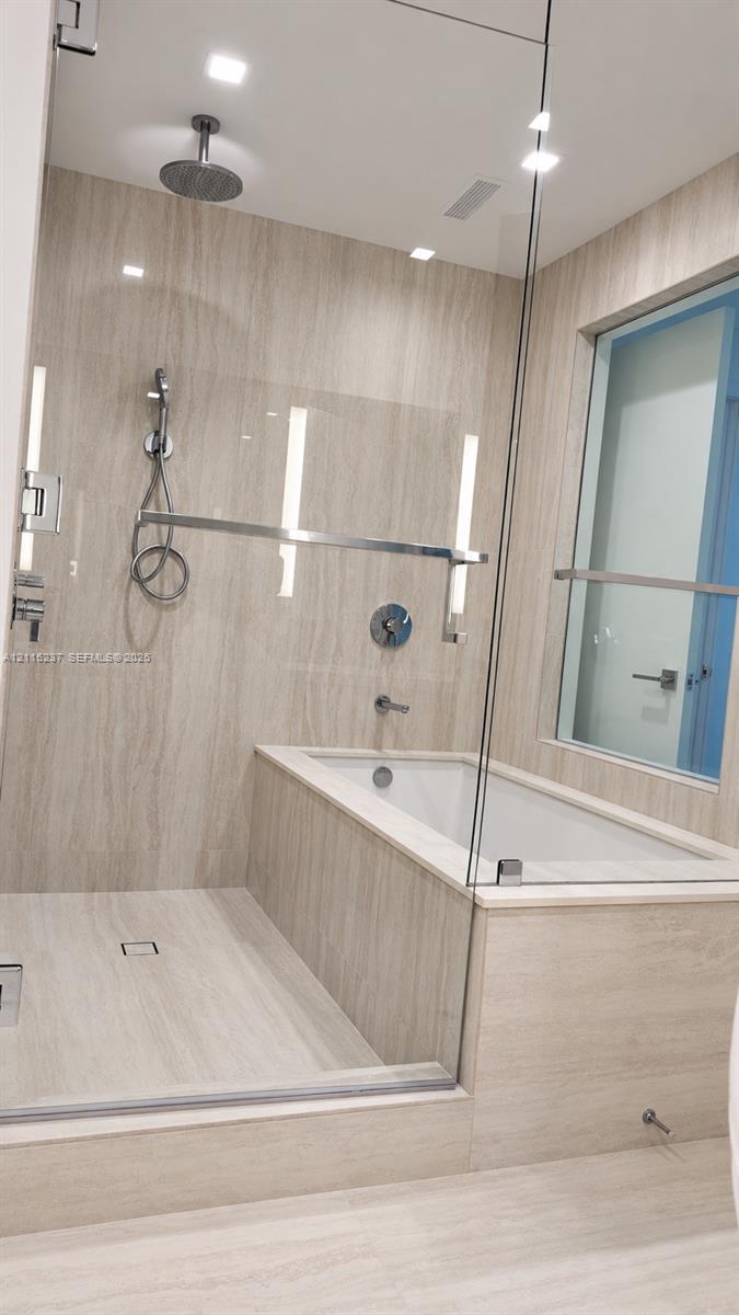 Glass-enclosed wet room featuring a built-in soaking tub, ceiling-mounted rainfall showerhead, handheld shower fixture, and floor-to-ceiling stone-finish tile surround