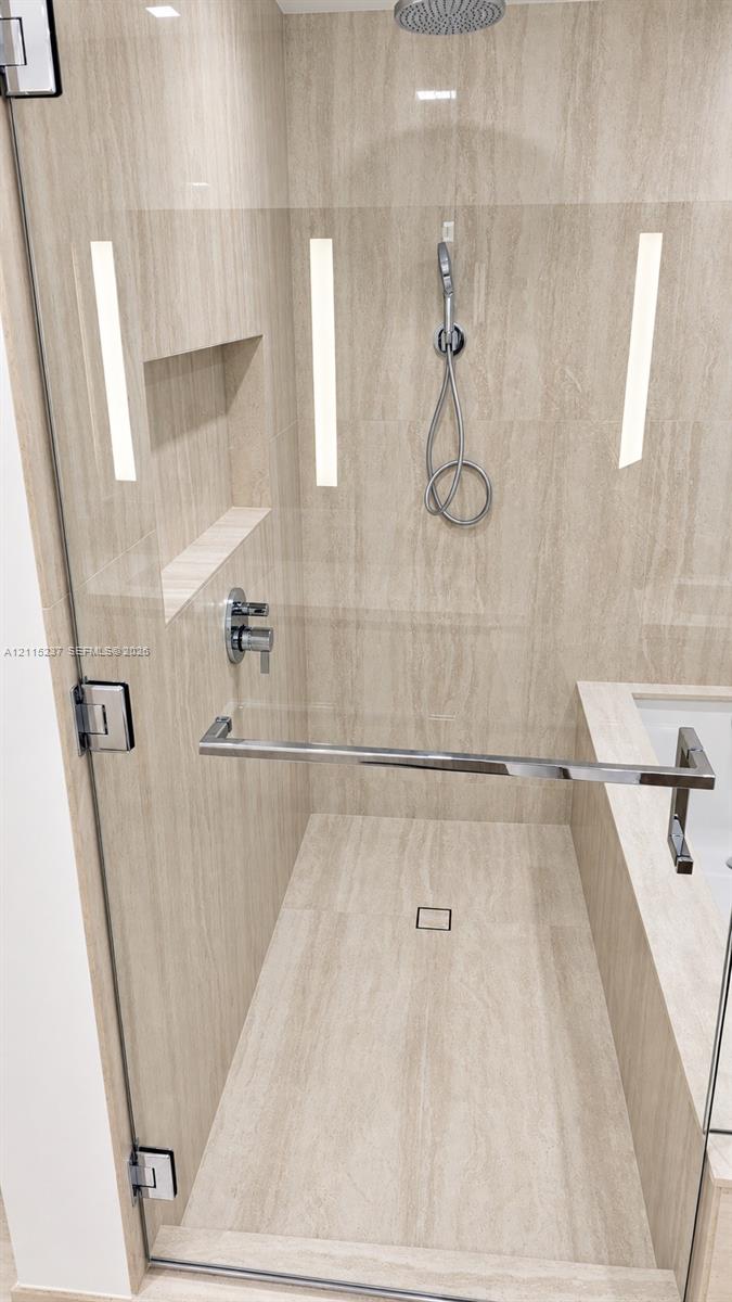 Walk-in shower featuring natural-stone tile surrounds, a rain-style showerhead with a handheld attachment, a built-in recessed niche, and a frameless glass enclosure with a metallic towel bar