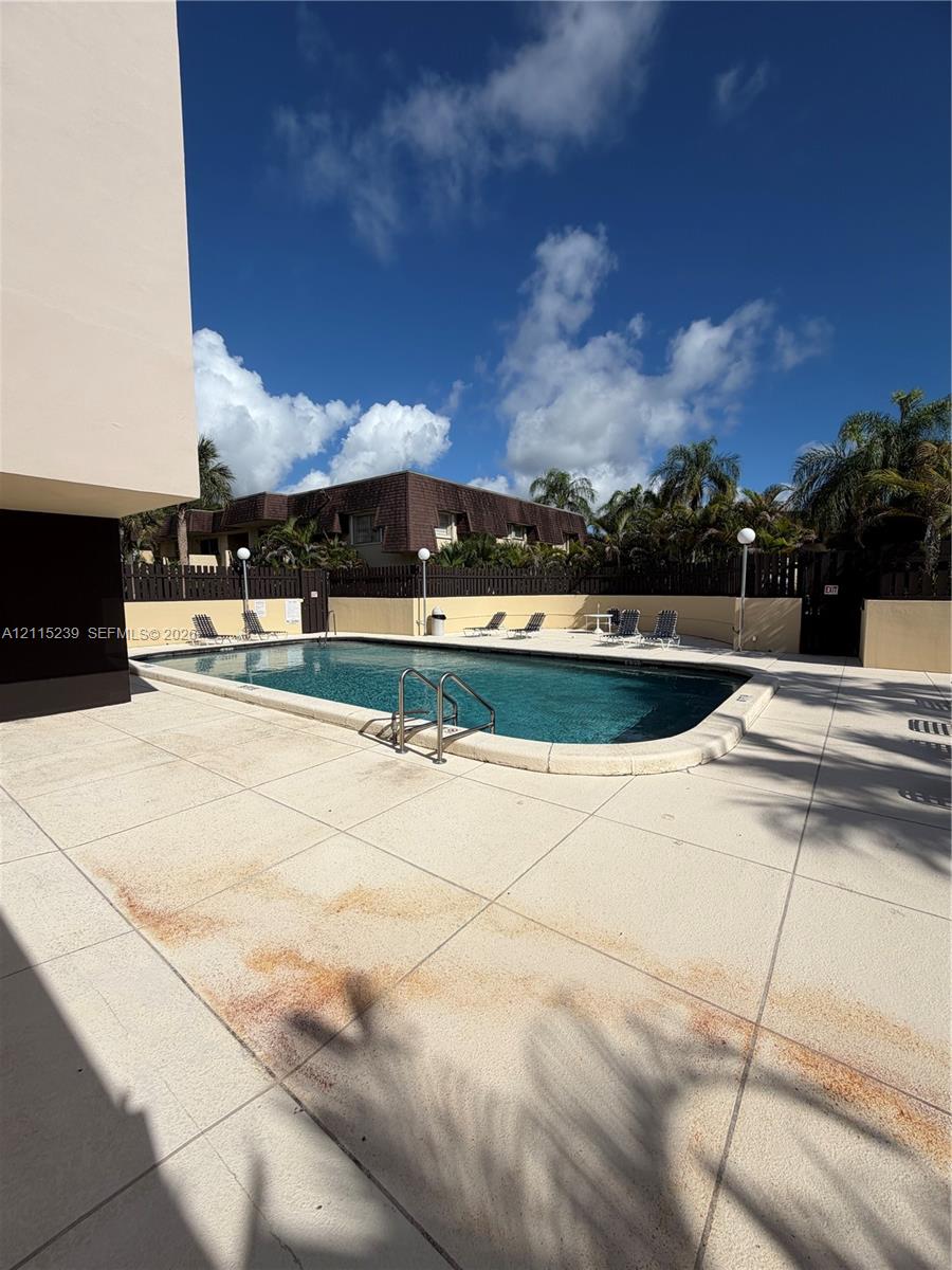 Outdoor swimming pool with rounded corners, metal handrail, concrete deck, perimeter fencing, and exterior lighting fixtures