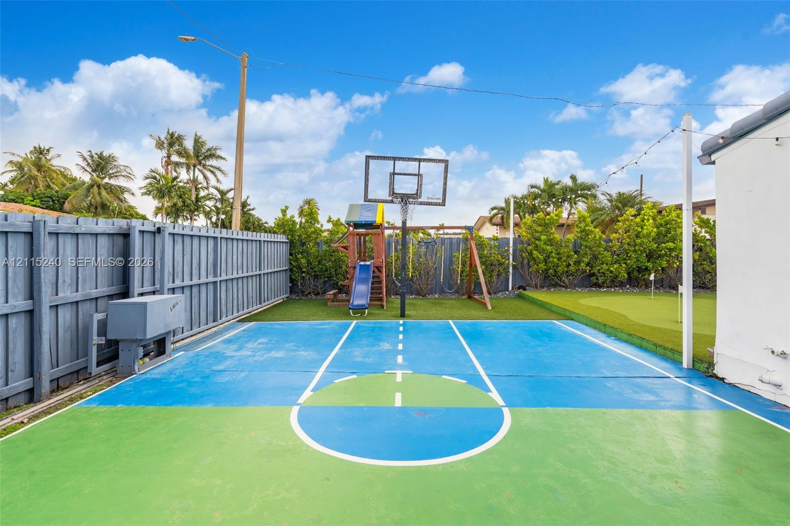 Outdoor area featuring a painted hard-surface court, a built-in basketball hoop, a dedicated putting green, and enclosed by wood-panel fencing