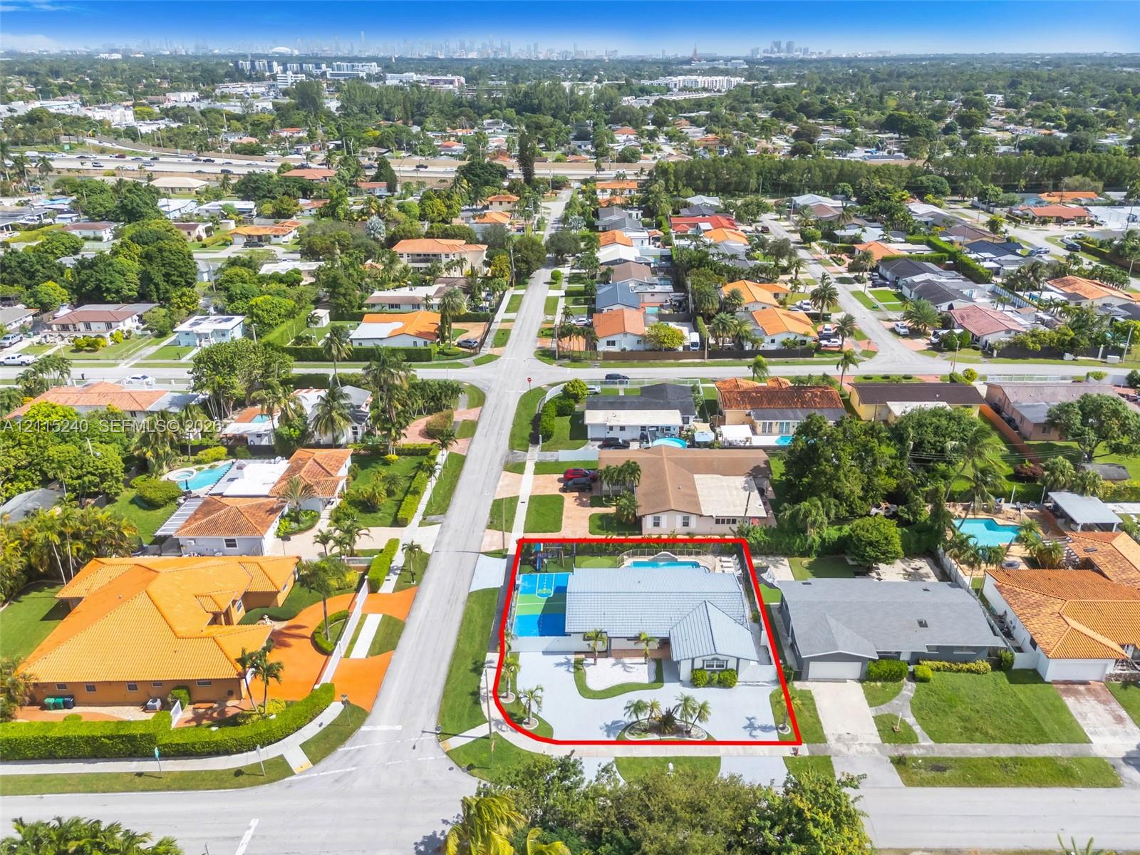 Aerial perspective of a corner lot property featuring a multi-pitched roof, an expansive paved driveway, a gated swimming pool, and a dedicated athletic court