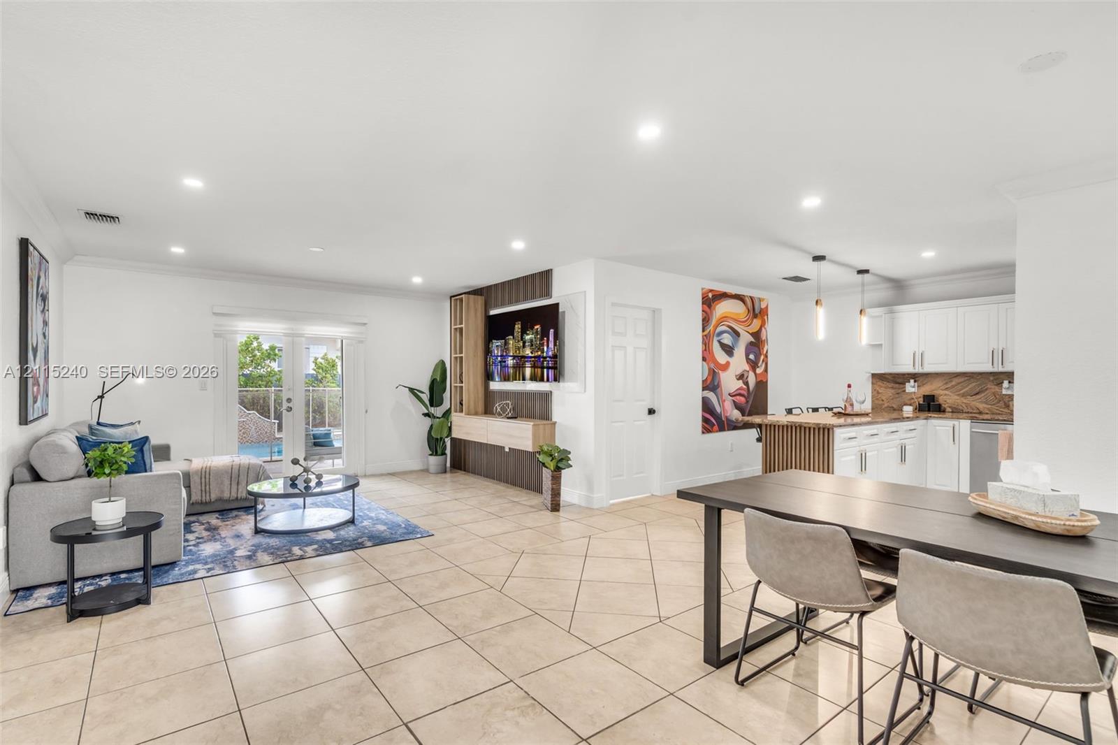 Open-concept interior featuring light-toned tile flooring, recessed ceiling lighting, a custom built-in media console with wood-slat detailing, white kitchen cabinetry with a textured backsplash, and double glass doors opening to an exterior area