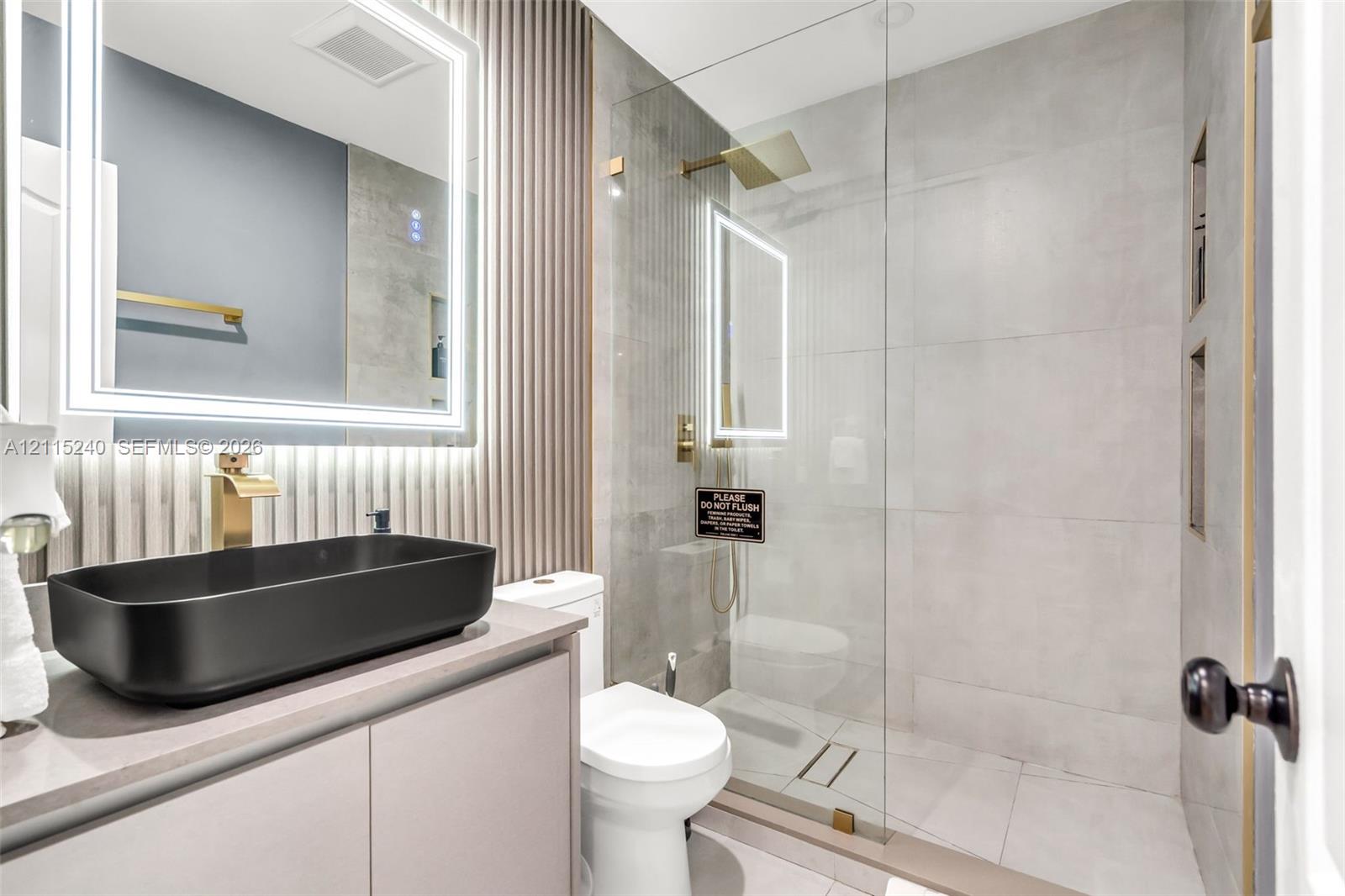 Bathroom featuring a matte black vessel sink, gold-toned faucet hardware, wall-mounted LED-backlit mirror, vertical slat accent wall, and a walk-in shower with large-format tile and a glass enclosure