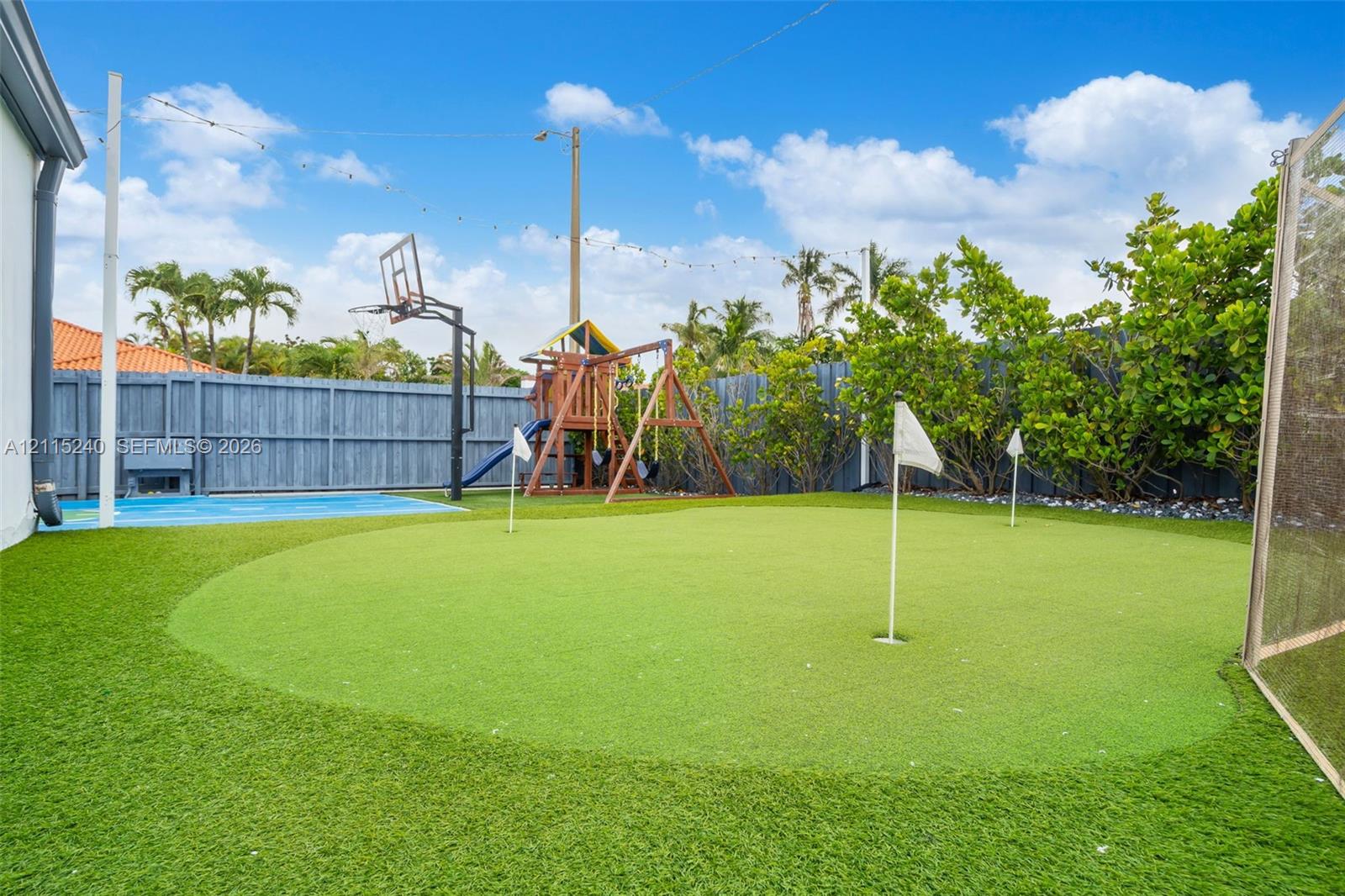 Backyard featuring a synthetic grass putting green, concrete court area, basketball hoop, and perimeter wood fencing