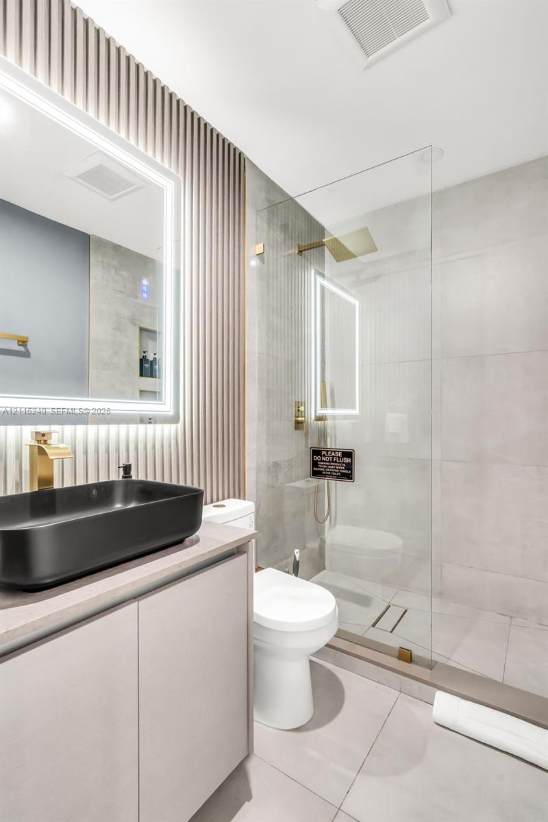 Bathroom featuring a slate-toned vessel sink, gold-tone hardware, a wall-mounted LED-backlit mirror, vertical slat accent wall, and a glass-enclosed walk-in shower with large-format tile