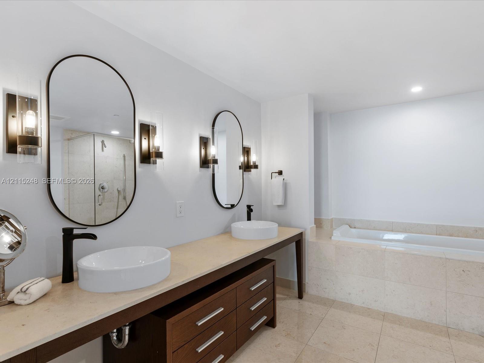 Dual vanity featuring stone-surfaced countertops, vessel sinks with matte black faucets, wood-finish cabinetry, and oval mirrors with integrated wall-mounted lighting, accompanied by a tiled soaking tub