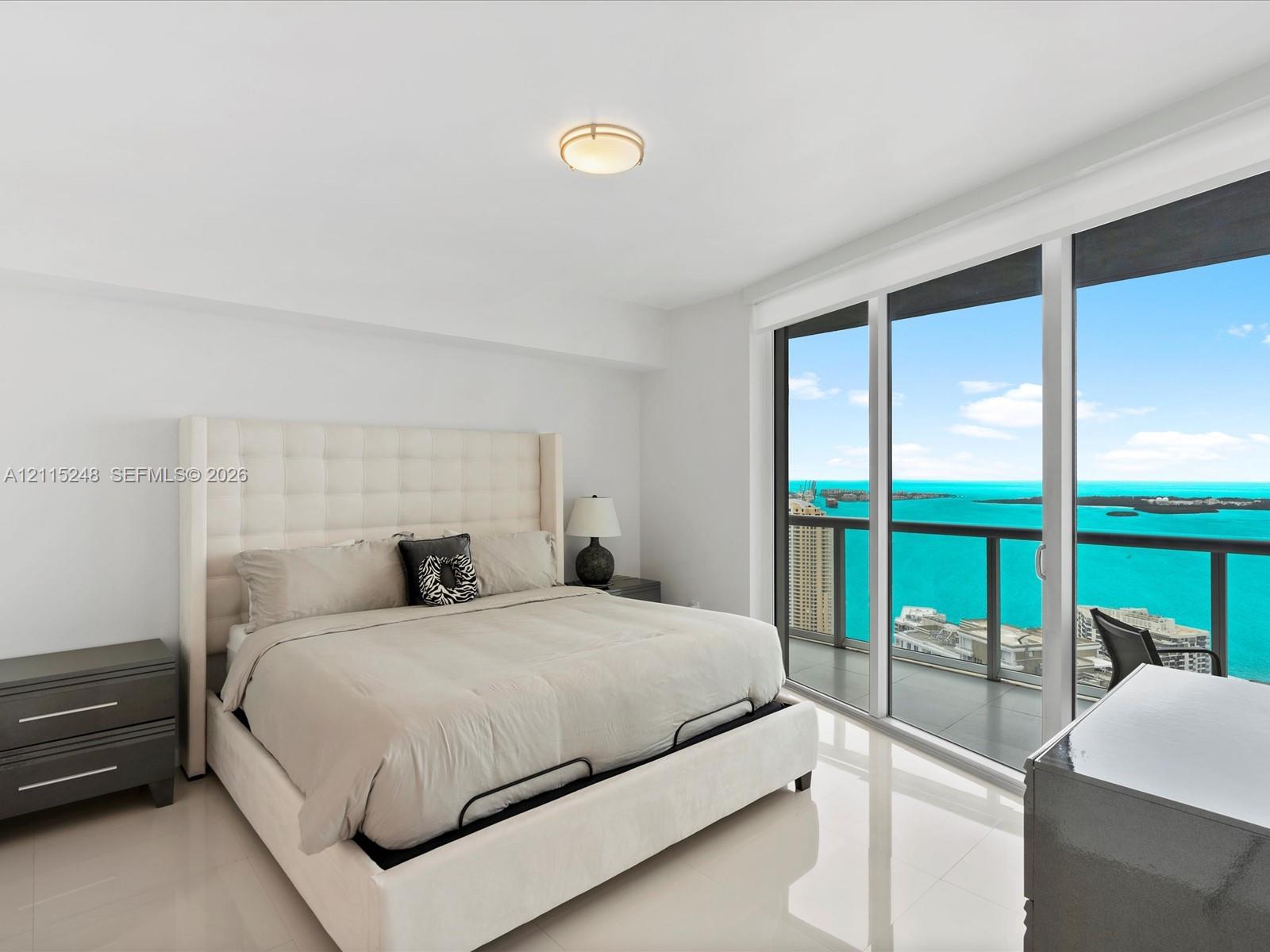Floor-to-ceiling glass sliding doors, private balcony with metal railing, neutral tile flooring, flush-mount circular lighting fixture, and panoramic water scenery