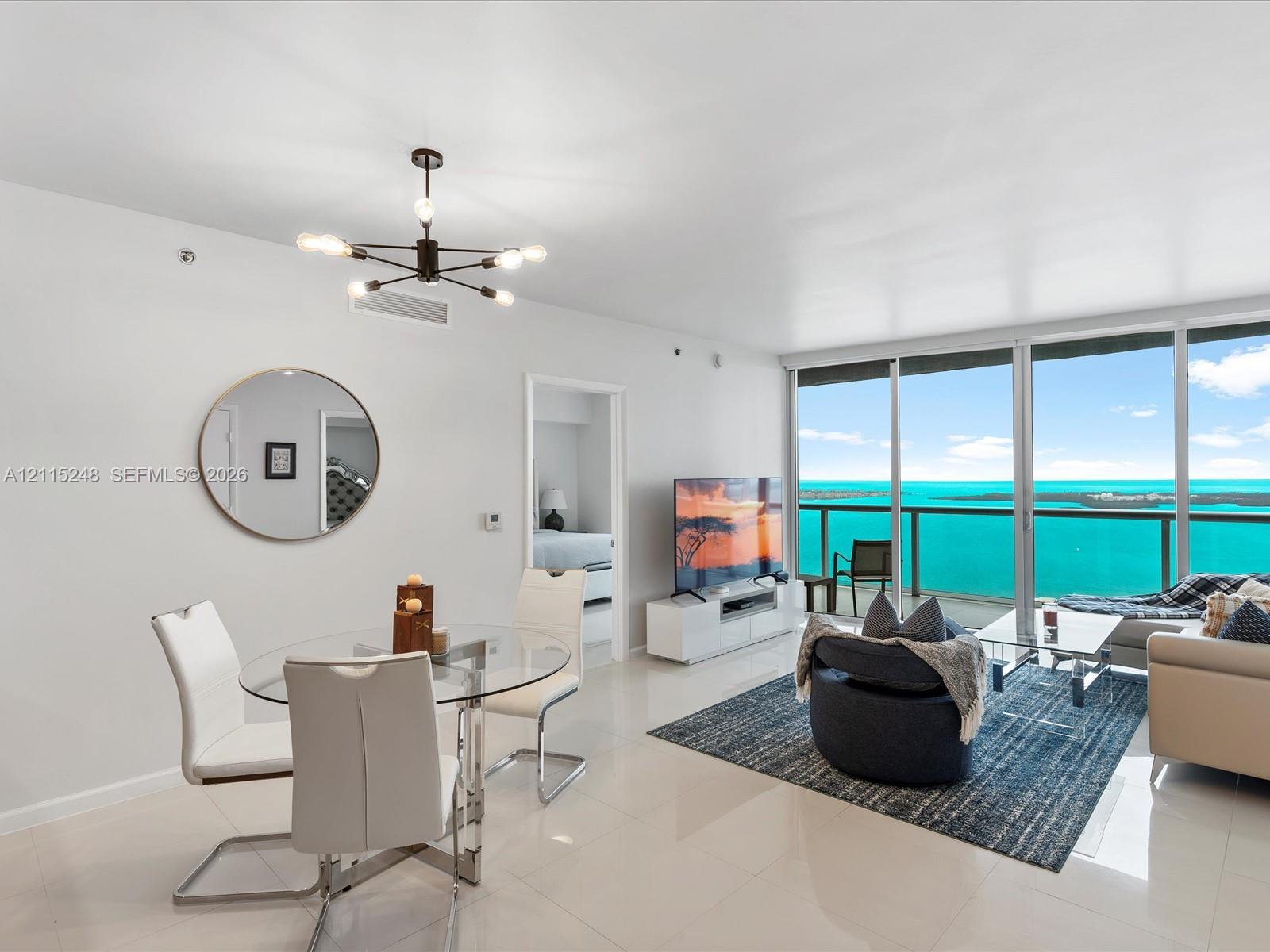 Open-concept space featuring floor-to-ceiling glass paneling, polished tile flooring, a geometric multi-arm pendant light fixture, and a glass-railed balcony overlooking the ocean