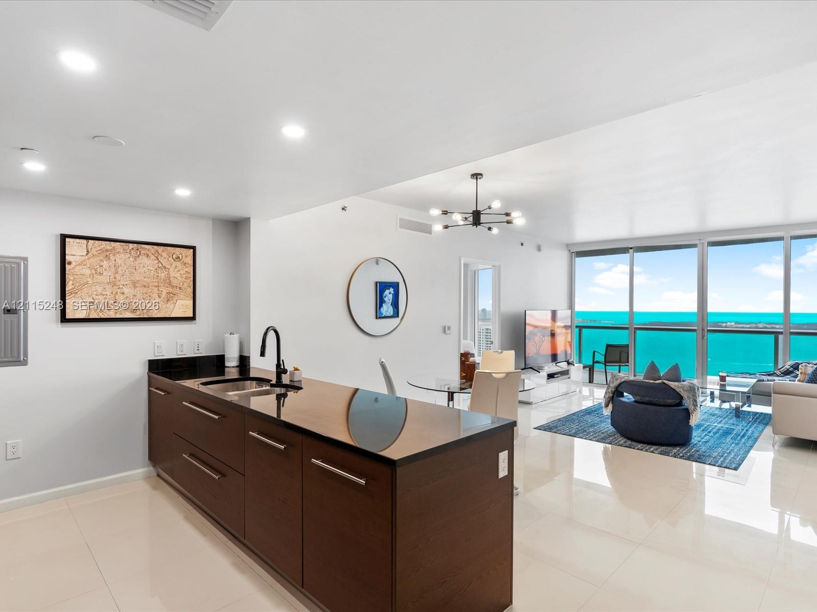Kitchen featuring a dark-toned island with integrated sink, matte black faucet, and polished stone-finish countertops, complemented by recessed lighting, a modern multi-arm chandelier, and floor-to-ceiling glass windows