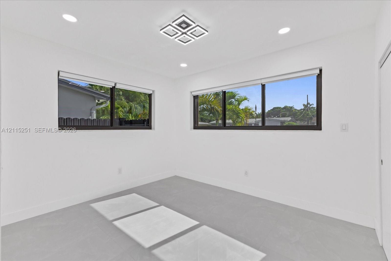 Interior room featuring neutral wall finishes, large dual-pane windows with dark frames, matte light gray tile flooring, recessed ceiling lighting, and a modern geometric flush-mount light fixture