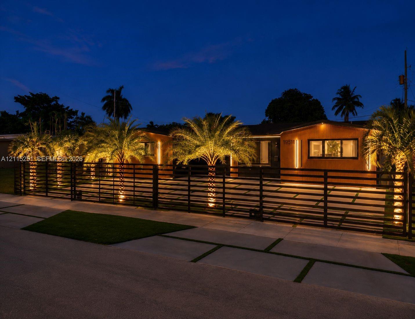 Single-story residence featuring horizontal wood-finish siding, a wide metal horizontal slat perimeter fence, paved driveway with grass grid accents, and mature palm trees Single-story residence featuring horizontal wood-finish siding, a wide metal horizontal slat perimeter fence, paved driveway with grass grid accents, and mature palm trees