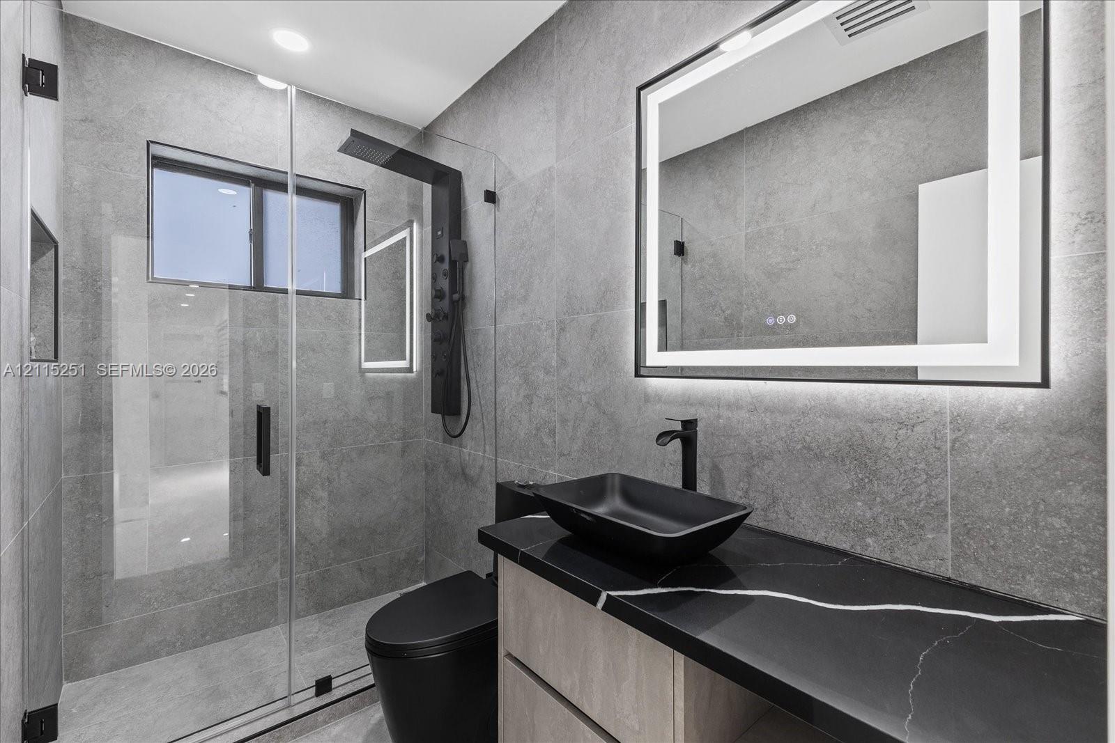 Bathroom featuring full-height large-format wall tile, glass-enclosed shower with matte black rainfall panel, floating vanity with black countertop, rectangular vessel sink, and illuminated vanity mirror