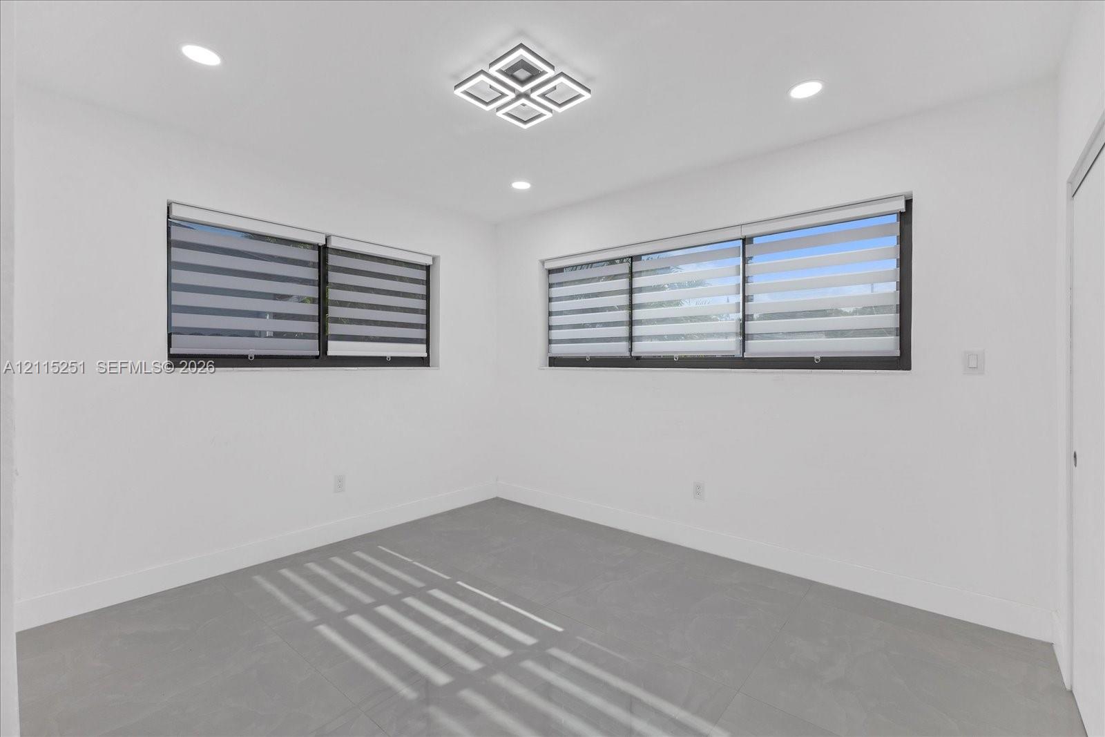 Neutral-toned room featuring large-format tile flooring, recessed ceiling lighting, a modern geometric flush-mount light fixture, and windows equipped with dual-layered roller shades