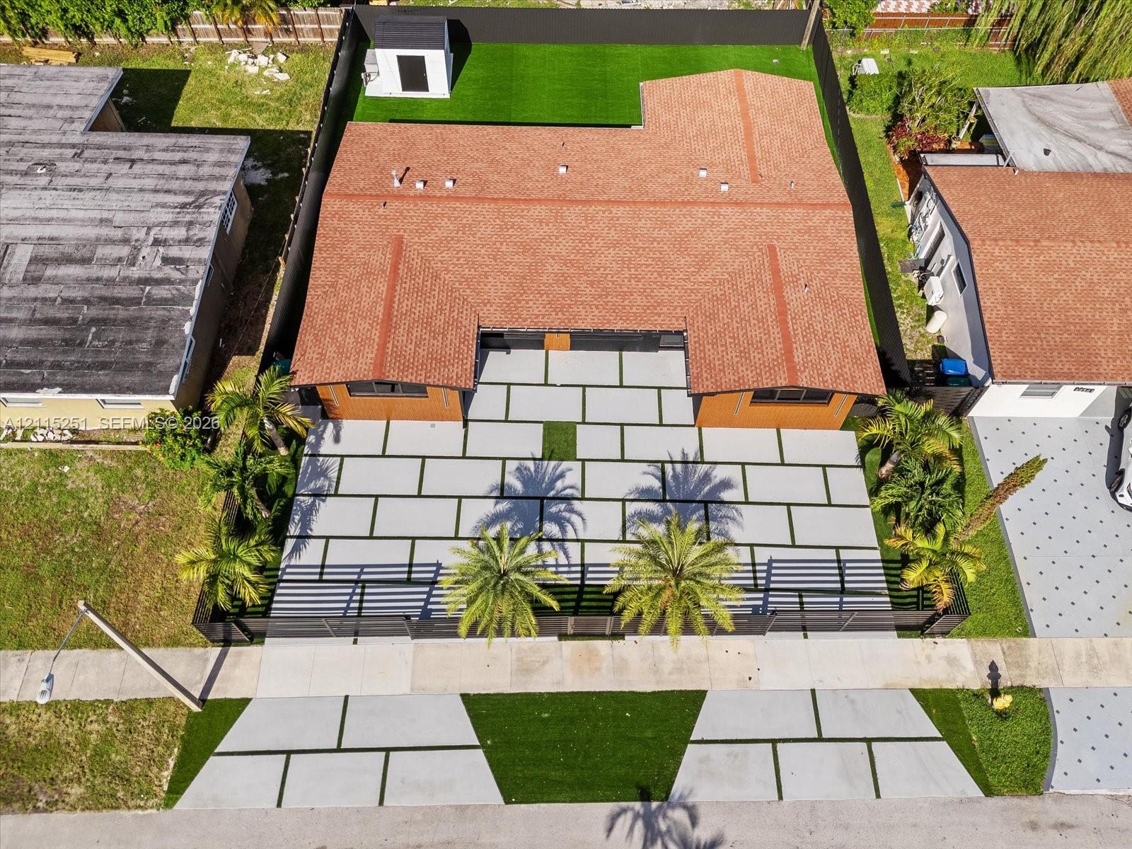 Aerial view of residential property featuring a large-format paver driveway, synthetic turf lawn, perimeter fencing, and mature palm trees