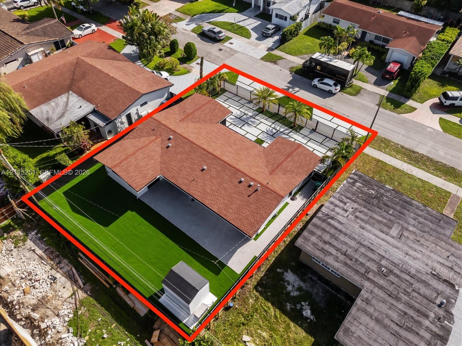 Aerial perspective featuring a U-shaped roofline, paved driveway with grass grid detail, synthetic turf lawn, perimeter fencing, and mature palm trees