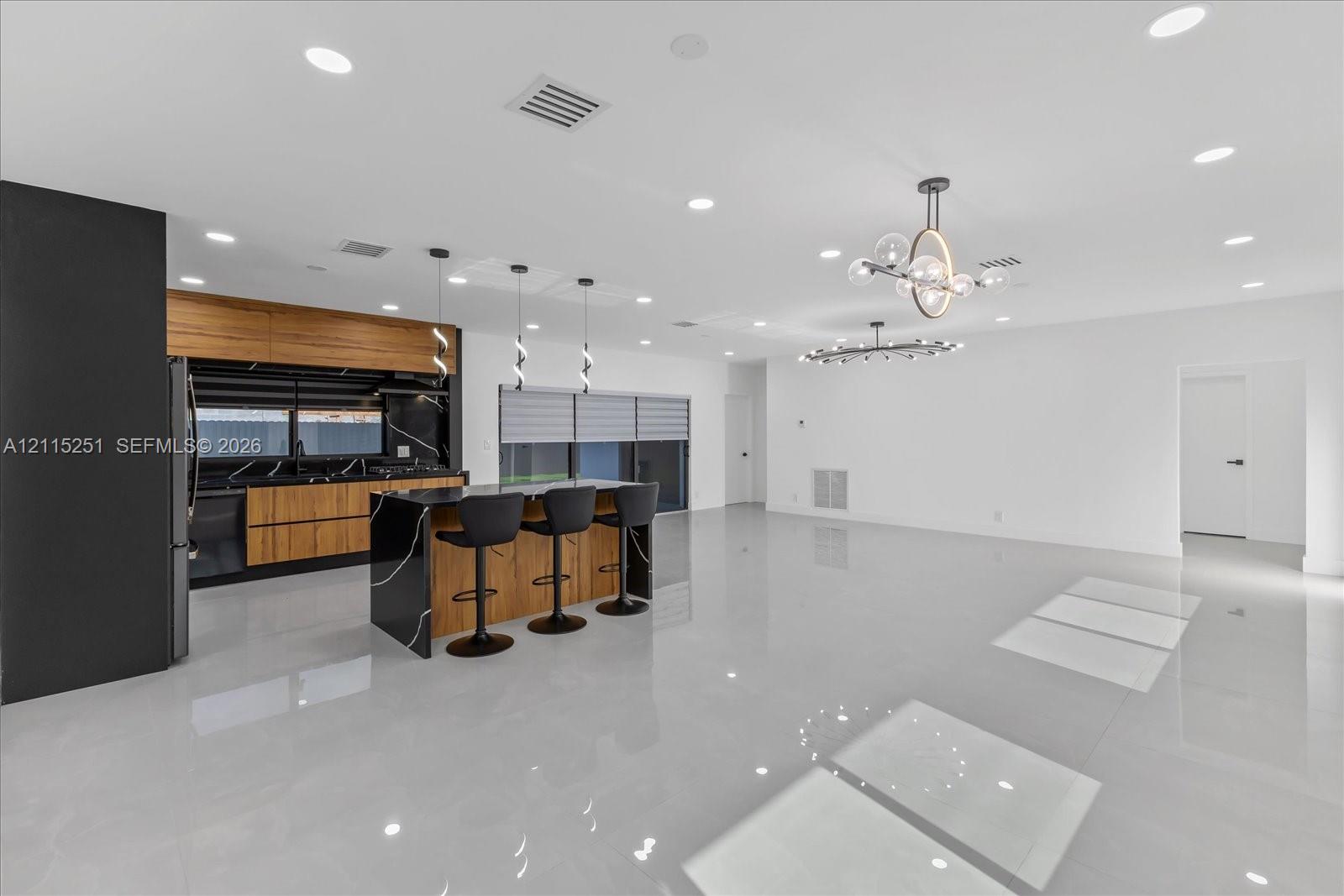 Kitchen featuring a large central island with waterfall-edge stone countertops, wood-finish cabinetry, black stone backsplash, recessed lighting, and modern multi-light chandeliers