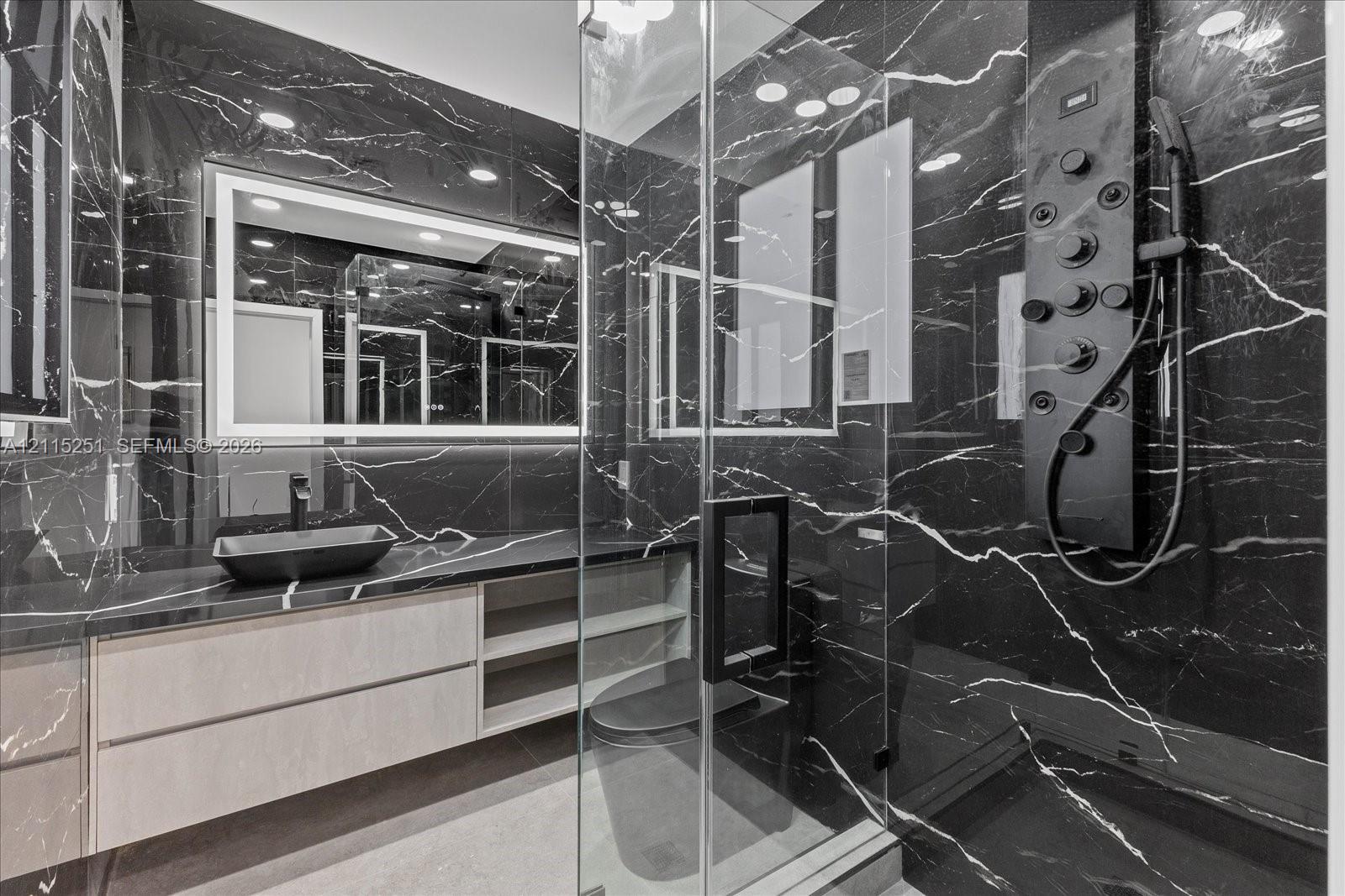 Floating vanity with integrated shelving, dark-veined stone surfacing, rectangular vessel sink, LED-backlit mirror, and glass-enclosed shower featuring a multi-functional matte black shower panel