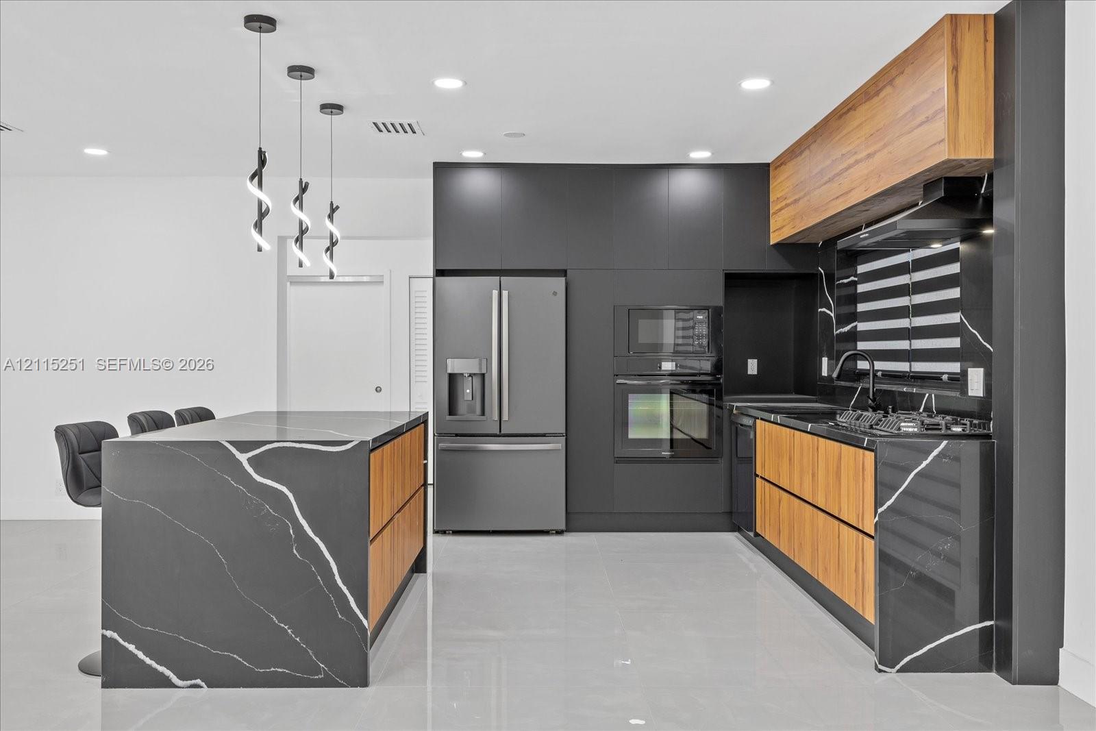Kitchen featuring a waterfall island with dark veined stone surfacing, flat-panel wood-finish cabinetry, built-in stainless steel appliances, matte black fixtures, and modern pendant lighting