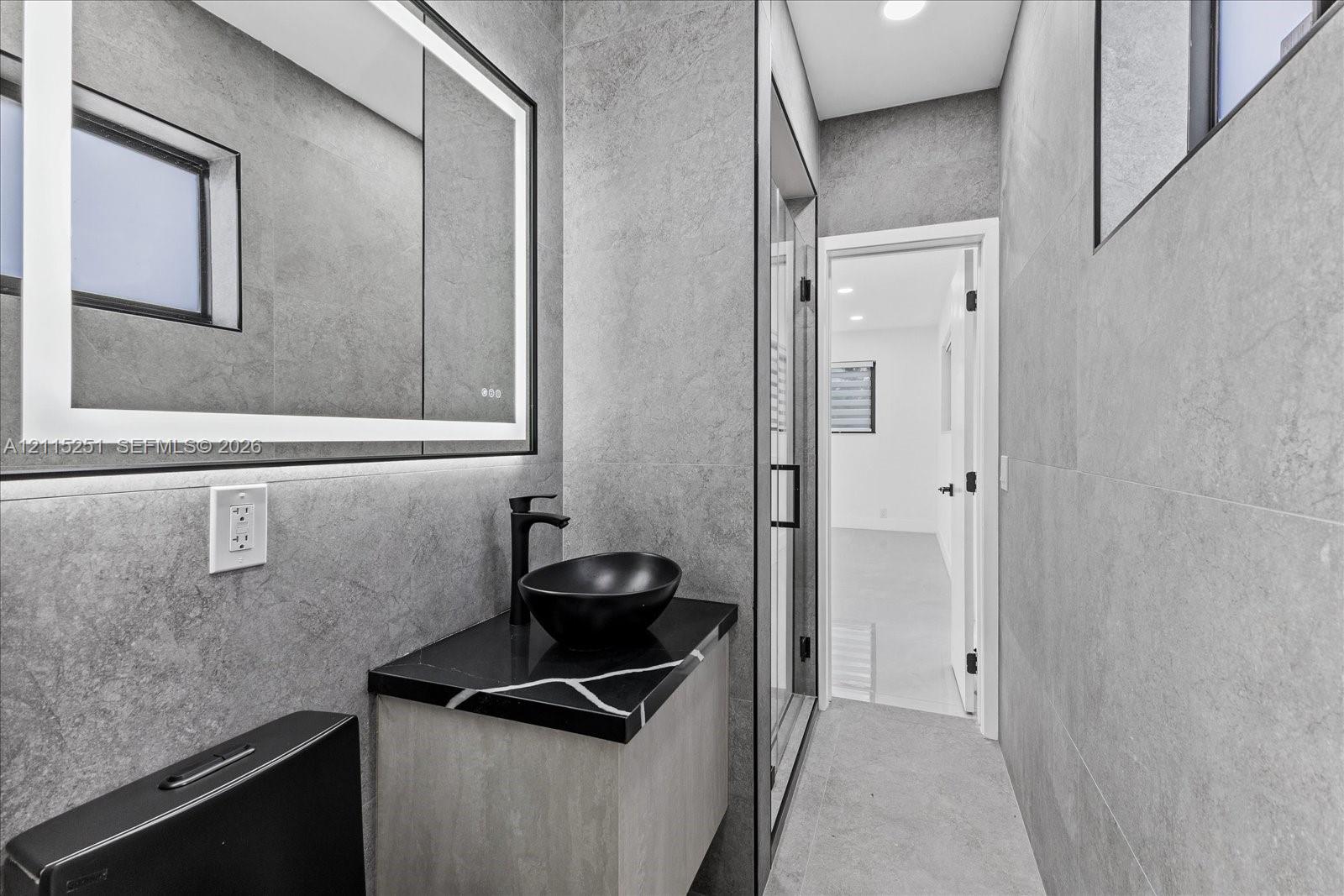 Modern vanity featuring a stone-finish countertop, black vessel sink, matte black faucet, integrated LED-backlit mirror, and floor-to-ceiling stone-style wall tile
